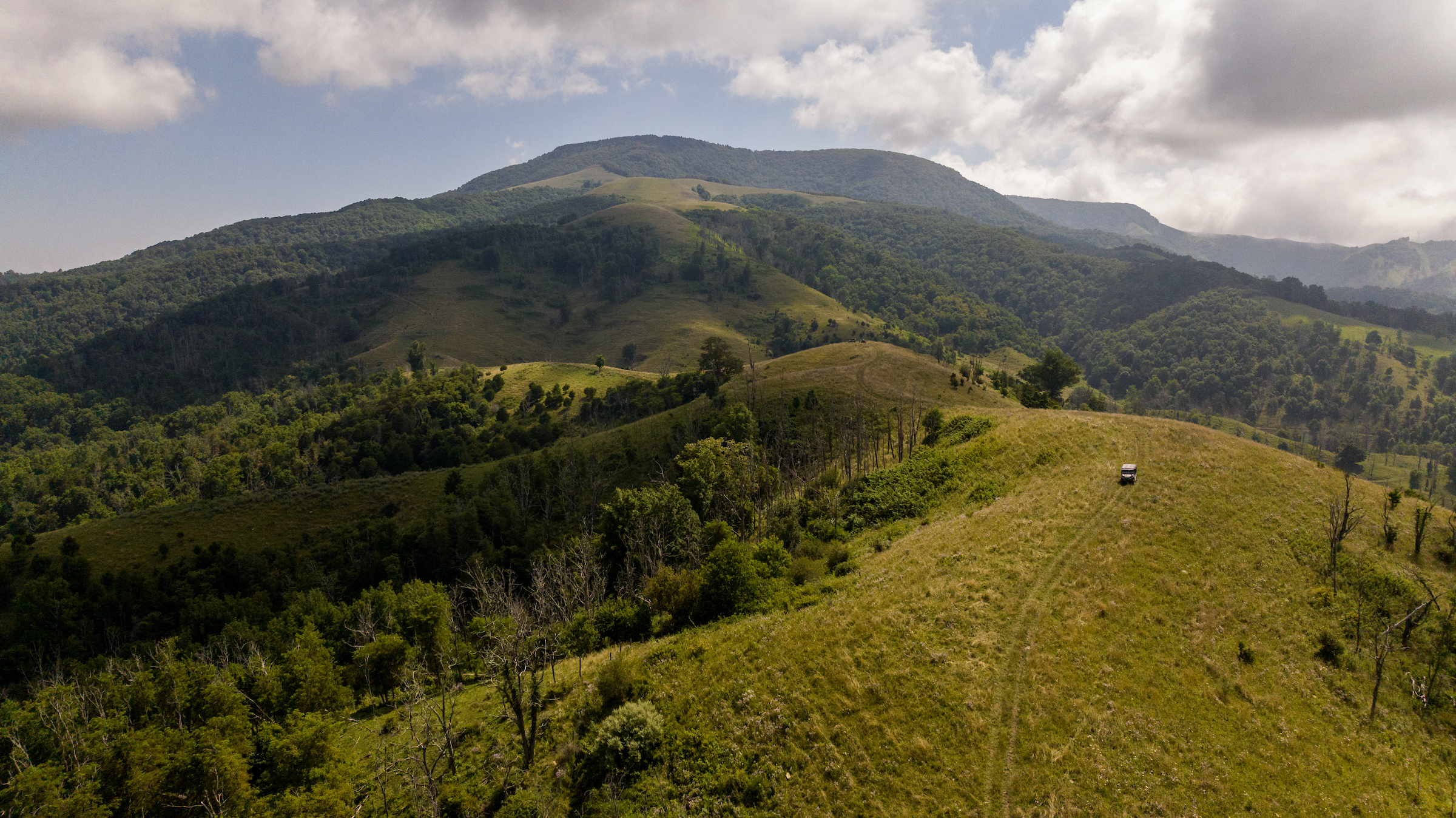 Aerial drone view of a large forested mountain ridge. A utility terrain vehicle drives along a curving, unpaved mountain track heading towards the summit.
