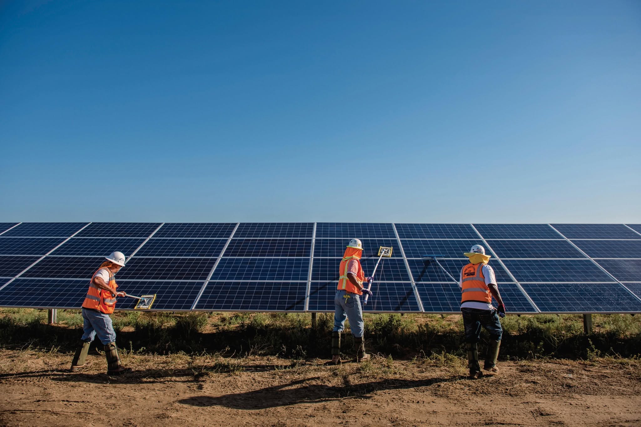 Three men in hard hats and orange utility vests clean large solar panels on a sunny day.