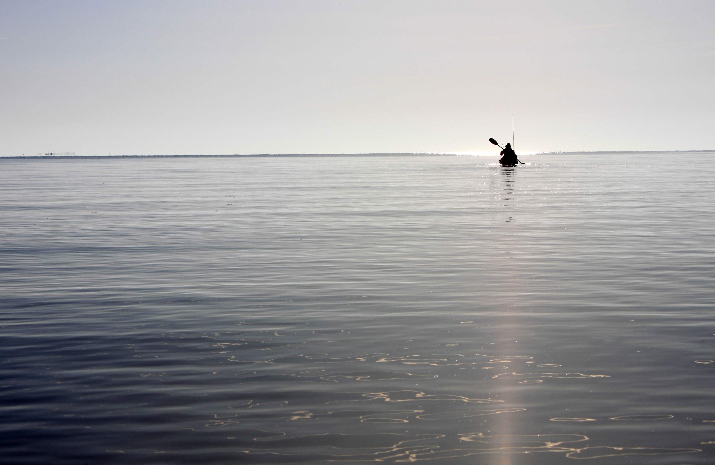 A person floats in a kayak in a flat, wide body of water. They are silhouetted against the sky and seem to hover in the distance at the horizon.