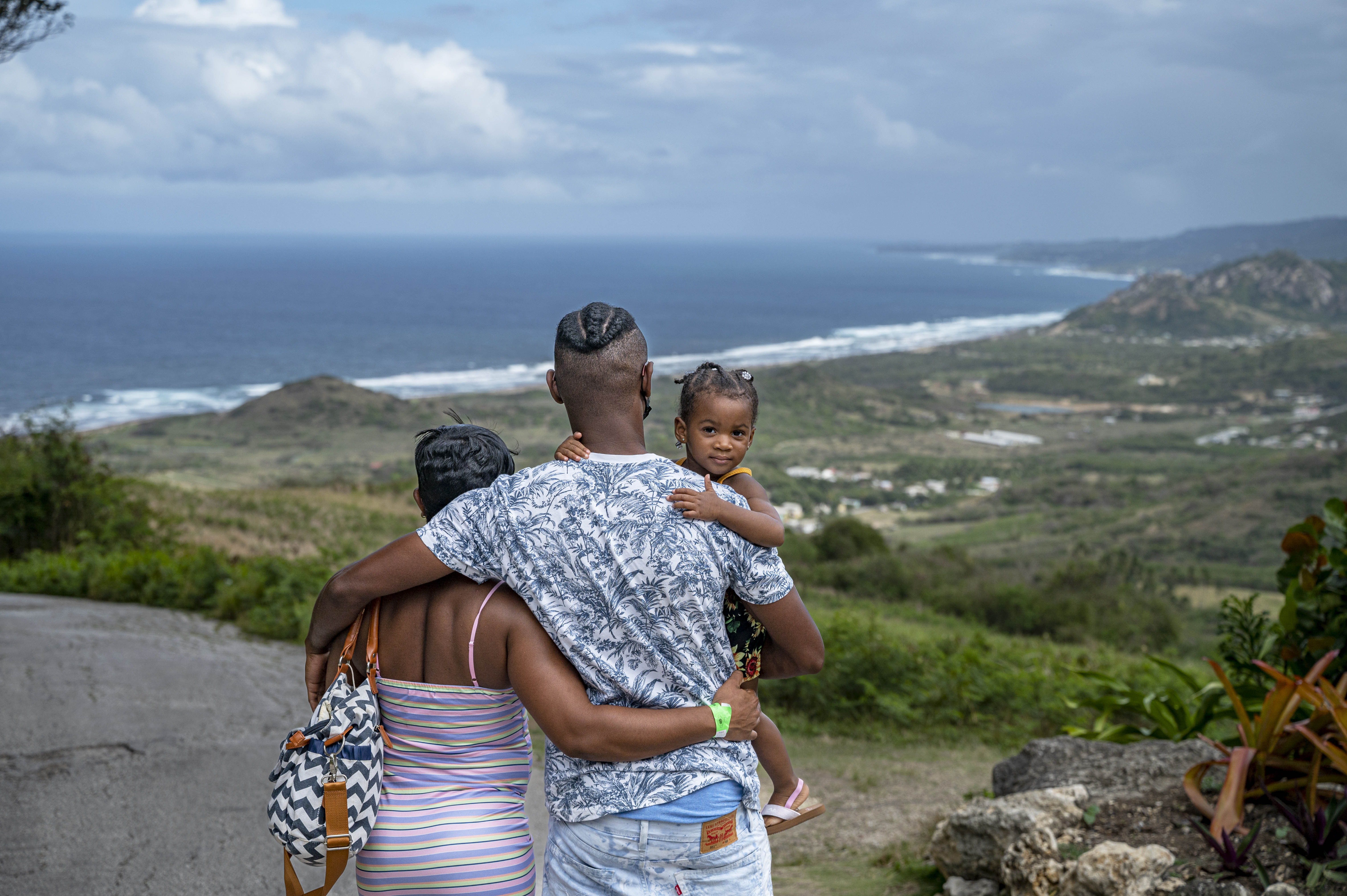 Photo of a family - father and mother holding young child - looking out over a Barbados hilltop toward the ocean.