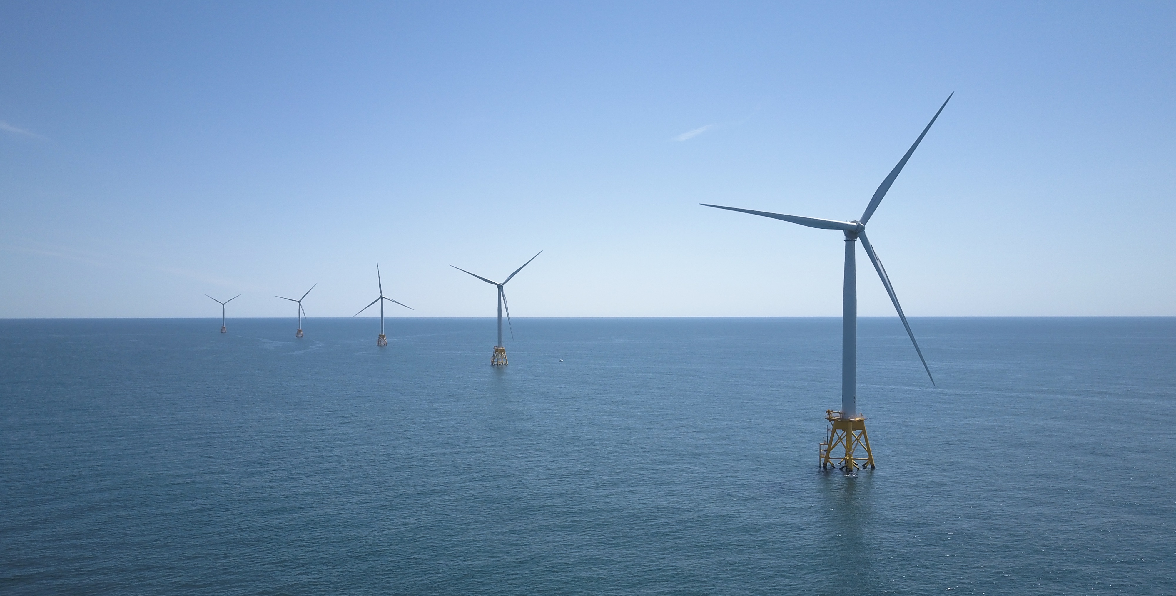 A view of five offshore wind energy turbines, with the right-hand turbine in the foreground stretching to the left-most turbine in the background. They sit on blue ocean water against a blue sky. 