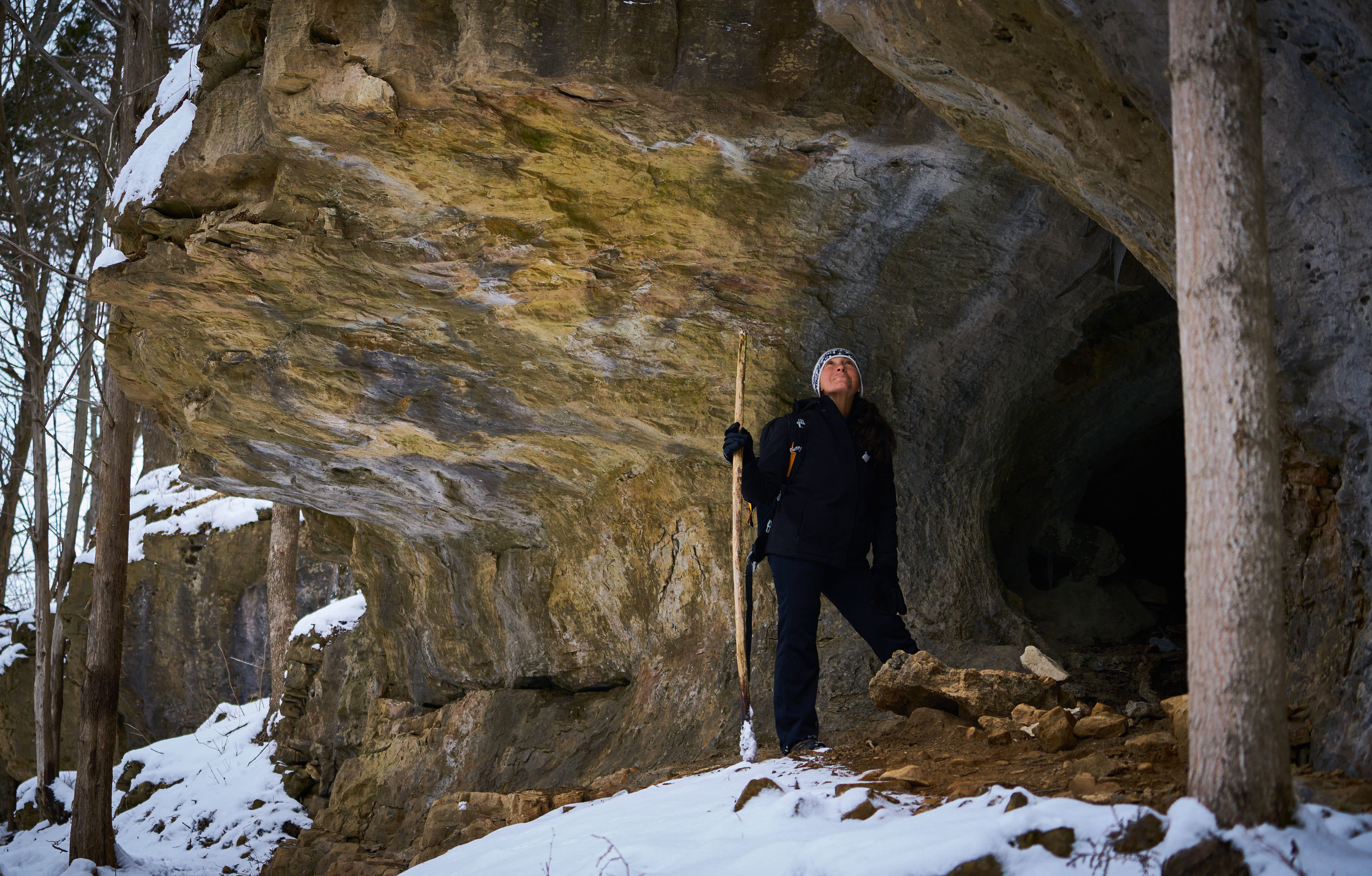 A hiker stands under a large rock on snow-covered ground.