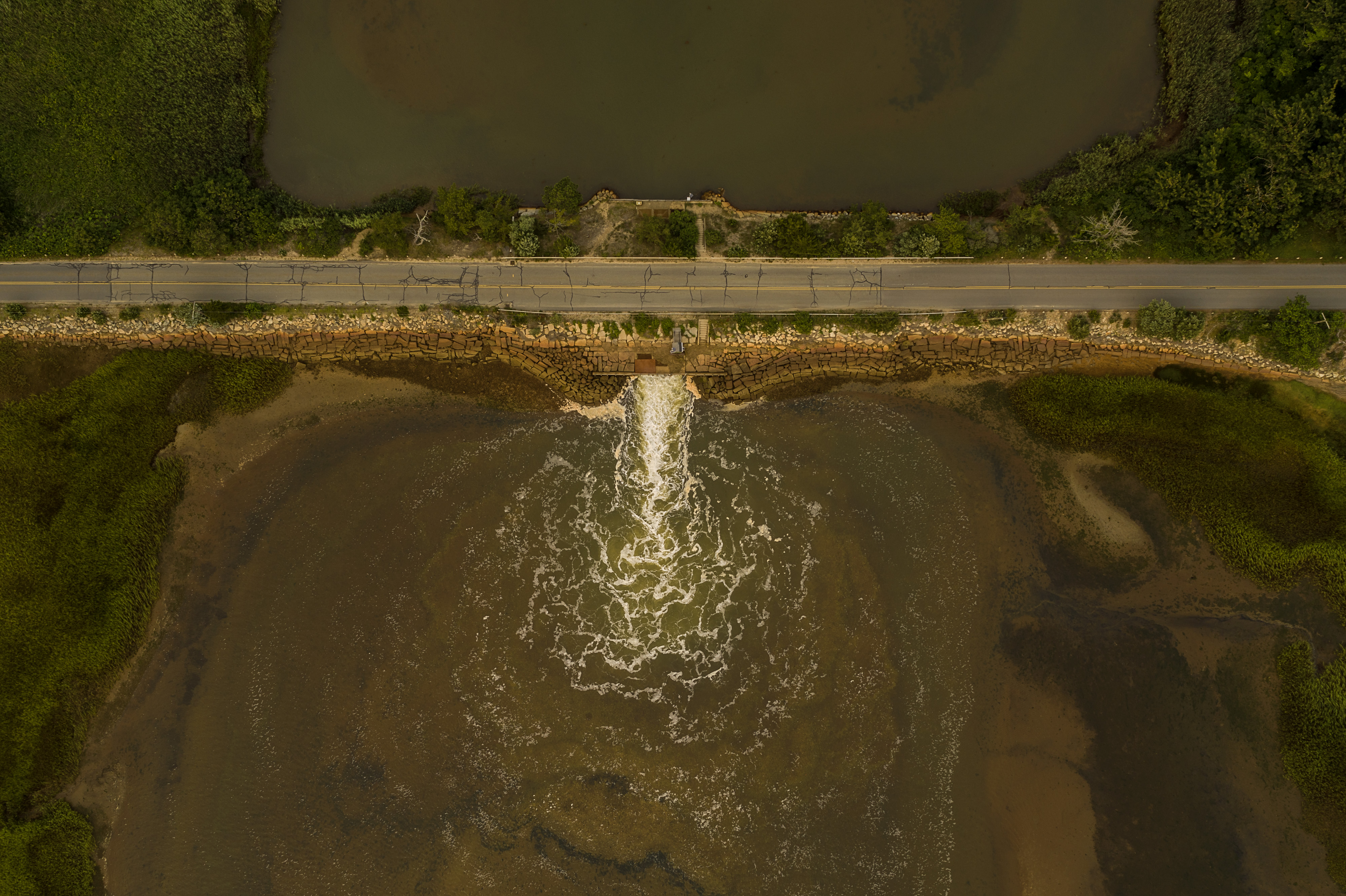 a cracked road stretching over a creek is seen from above with water spilling out of a narrow hole on one side