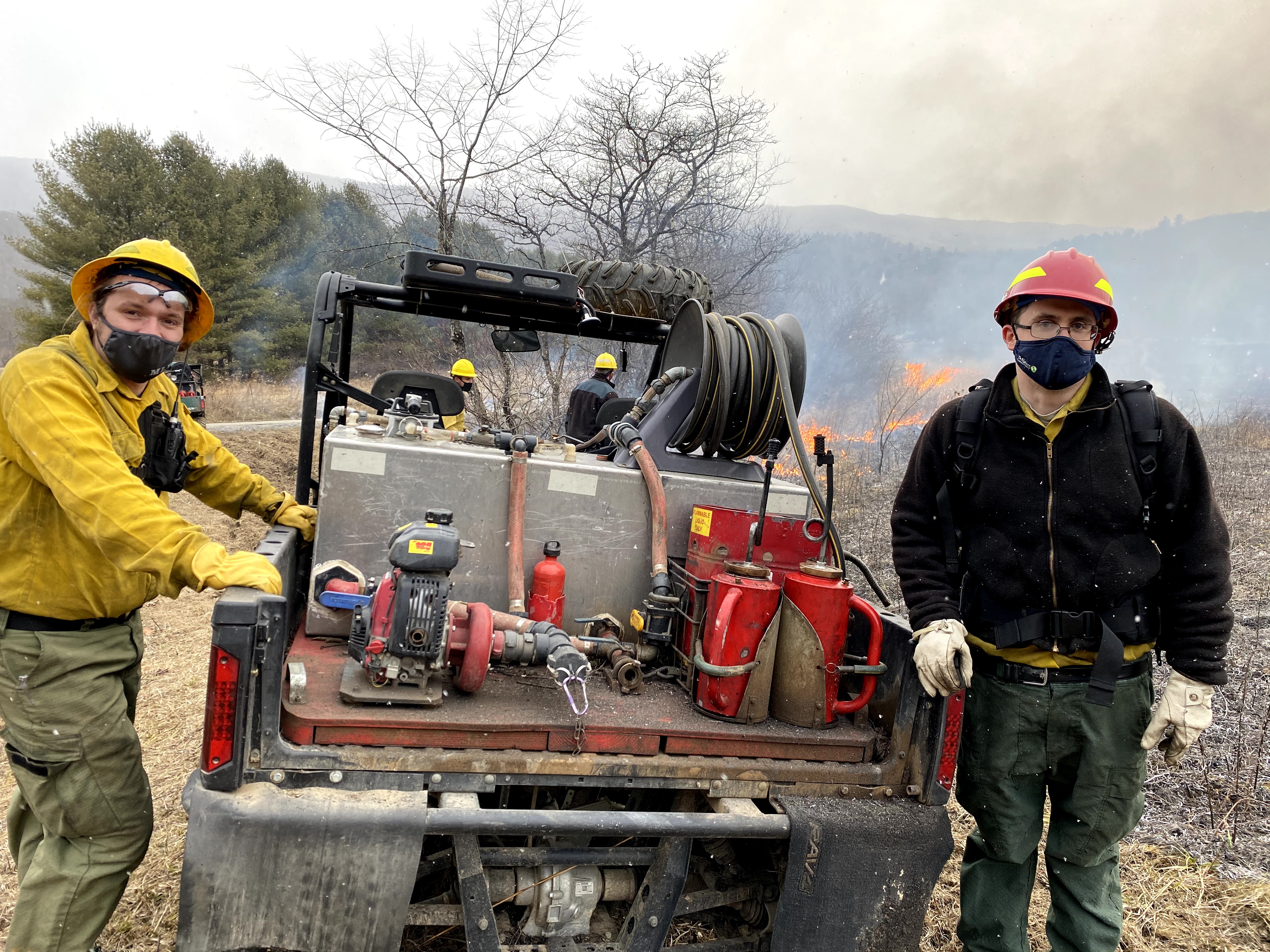 Two men wearing fire gear stand on either side of a small fire engine. Behind them two men monitor a fire during a controlled burn. The air is filled with flecks of either snow or ash.