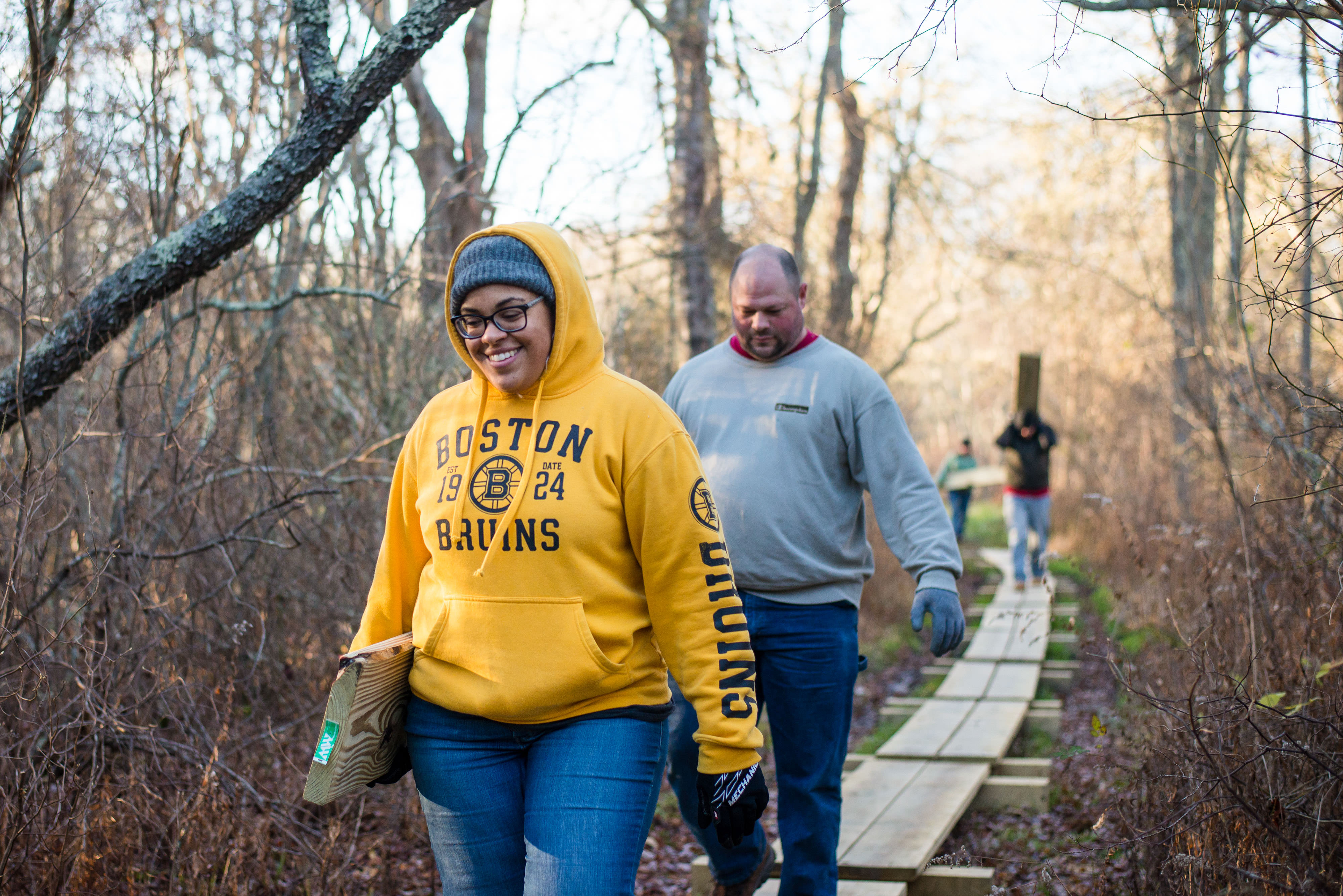 Several people walk along a boardwalk toward the camera in a wooded area.