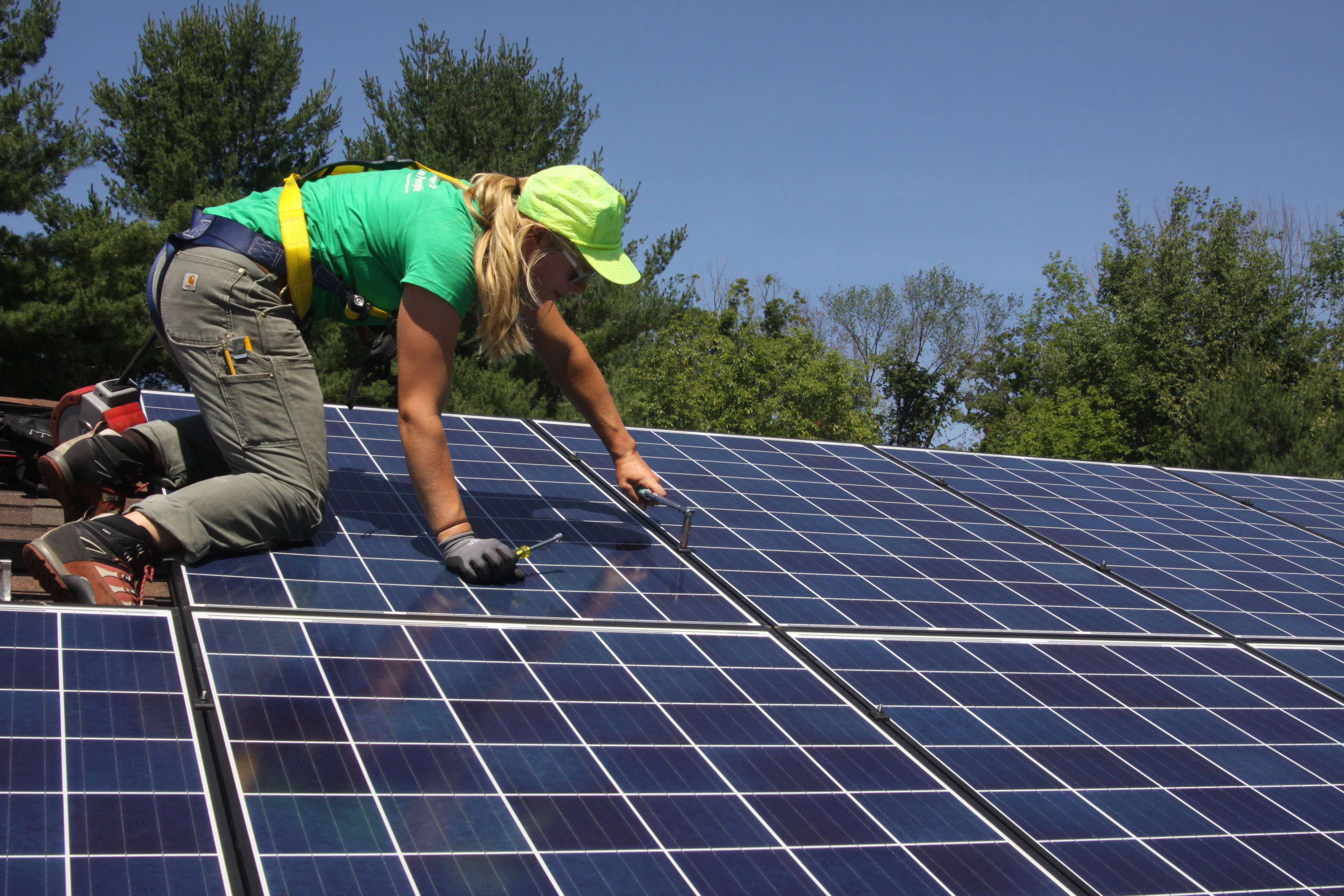A solar energy installer working on a solar panel with green trees in the background.