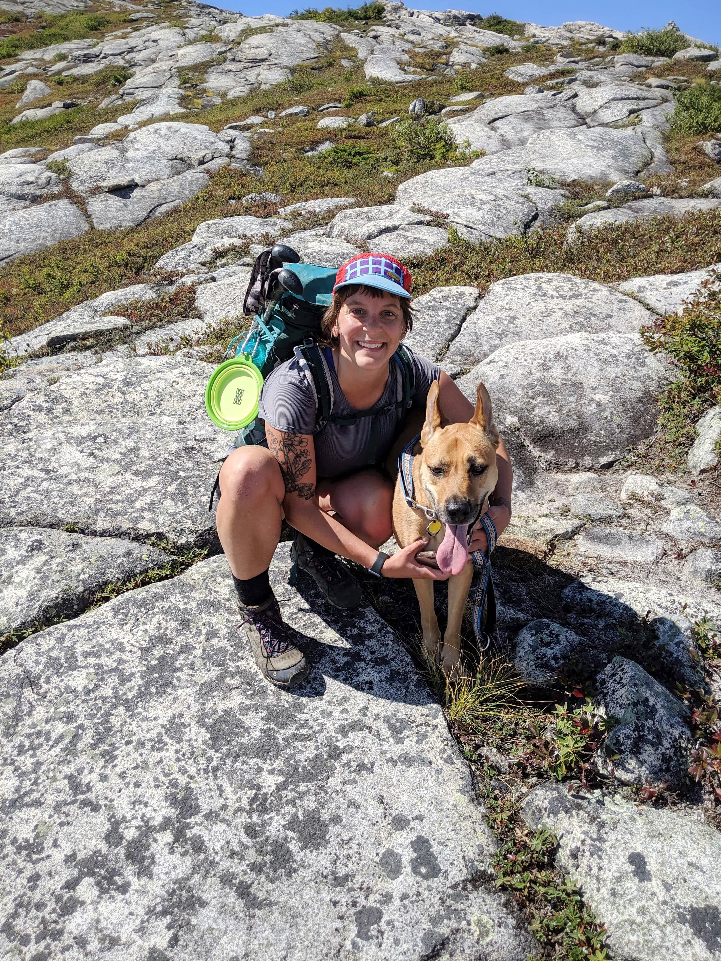 A woman in hiking gear kneeling on a rocky mountain top next to her brown dog.
