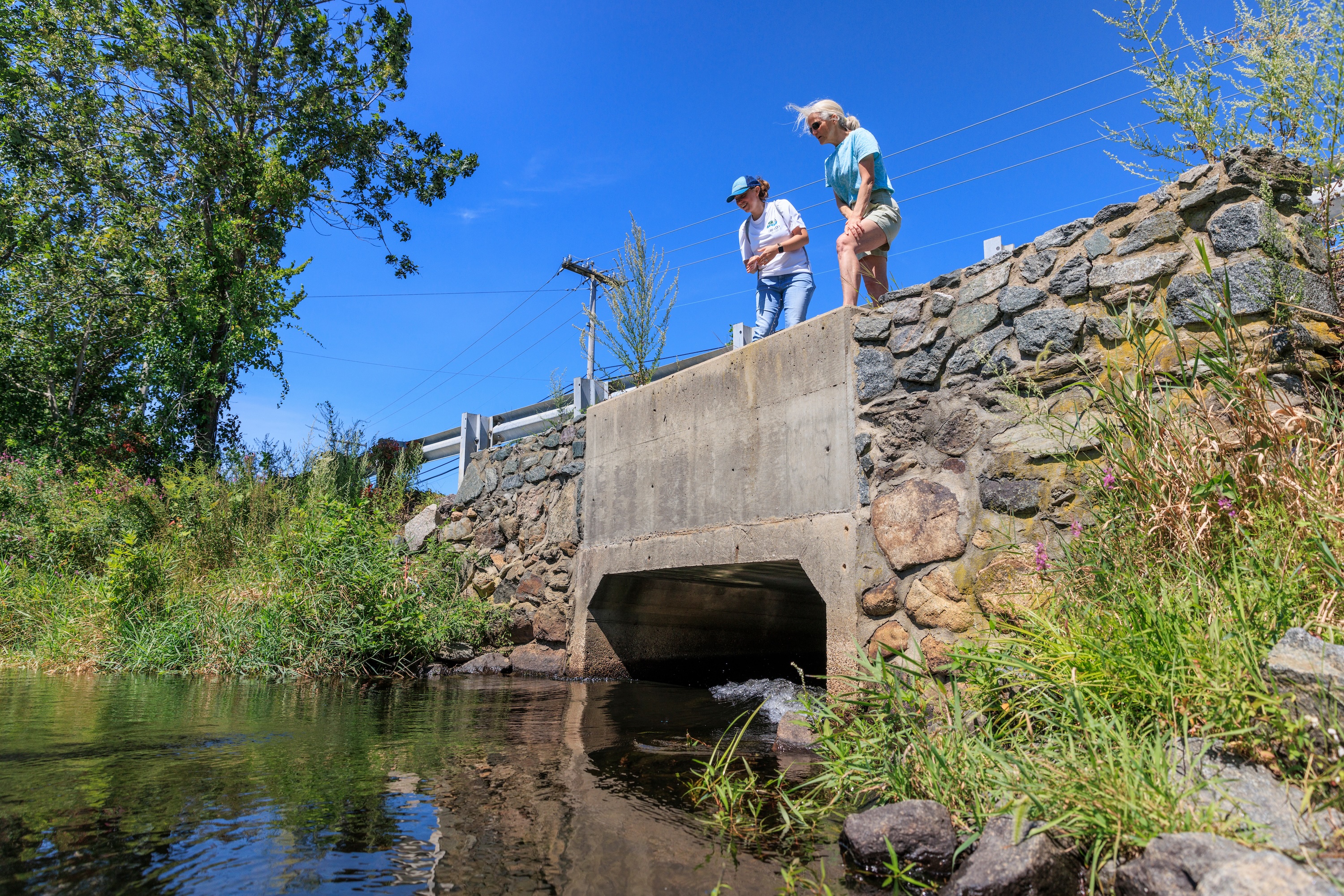 Two woman stand above a cement and stone culvert passing under a road in Lakeville, MA, that connects Assawompset Pond with Snake River.