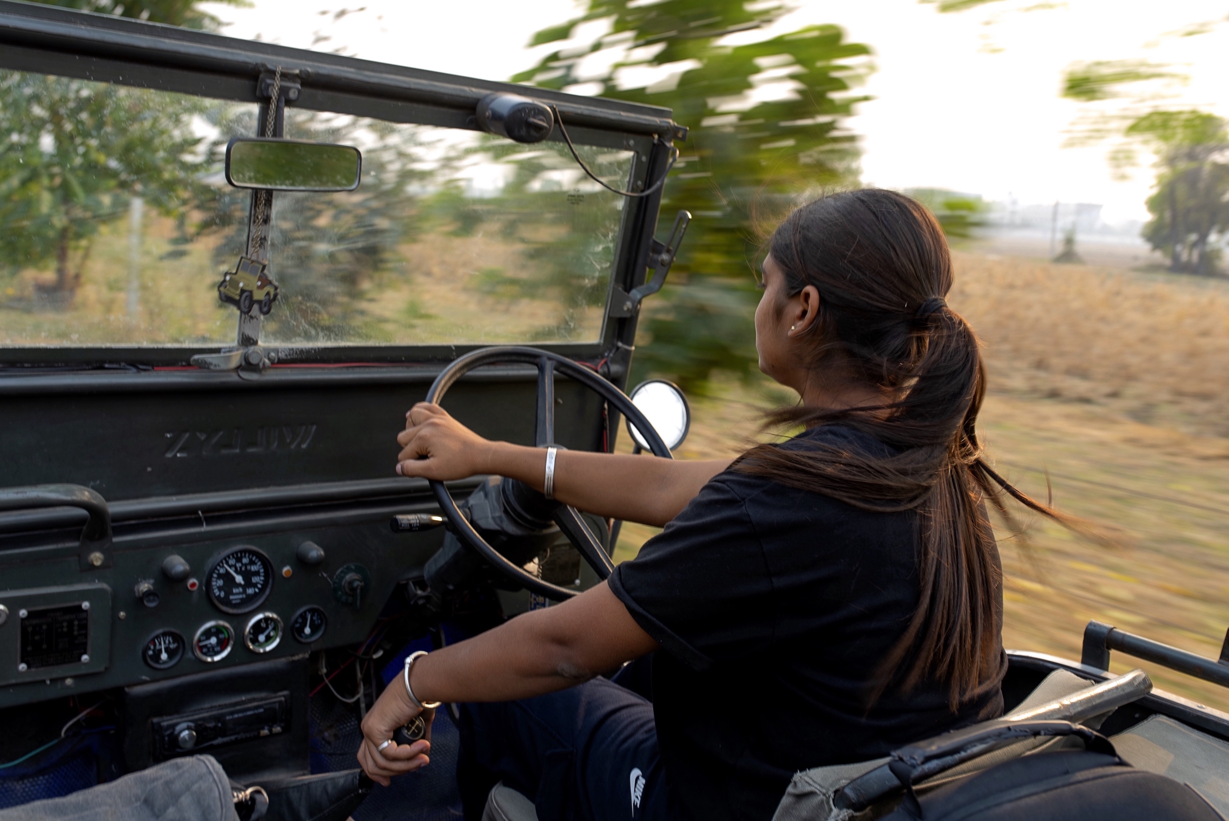 A view from the passenger side of an open jeep as Amandeep Kaur drives through a farming region, the landscape flying in motion around her.