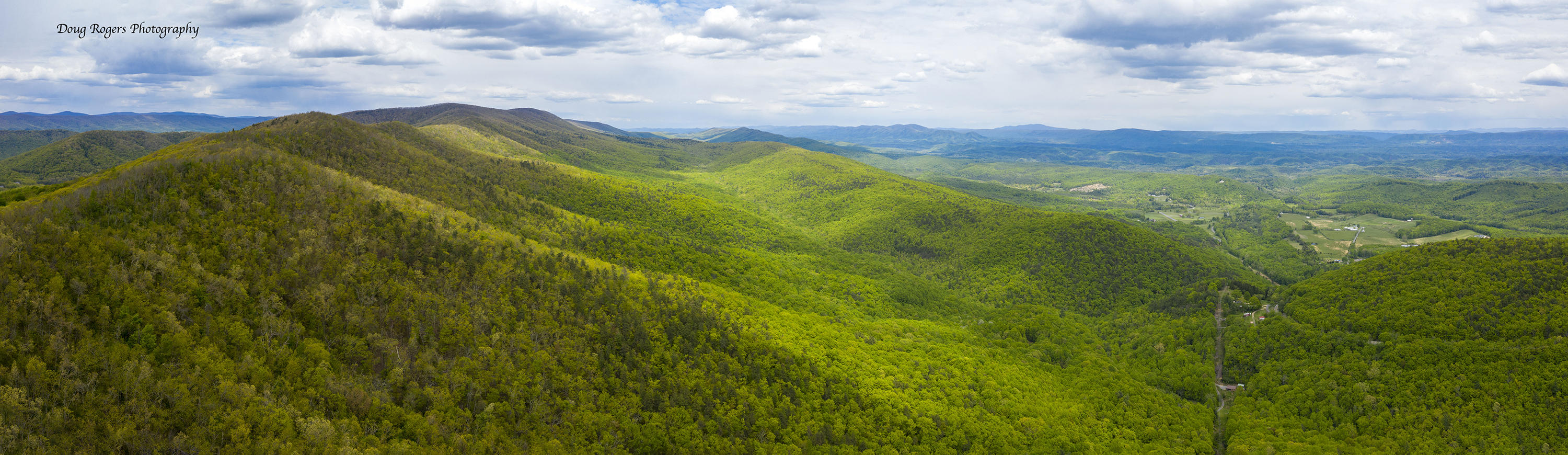 Drone view looking out over rolling mountain ridges and valleys. The mountain sides are thickly forested. Sunlight dapples over the trees. Small clearings and farms are visible in the distance.
