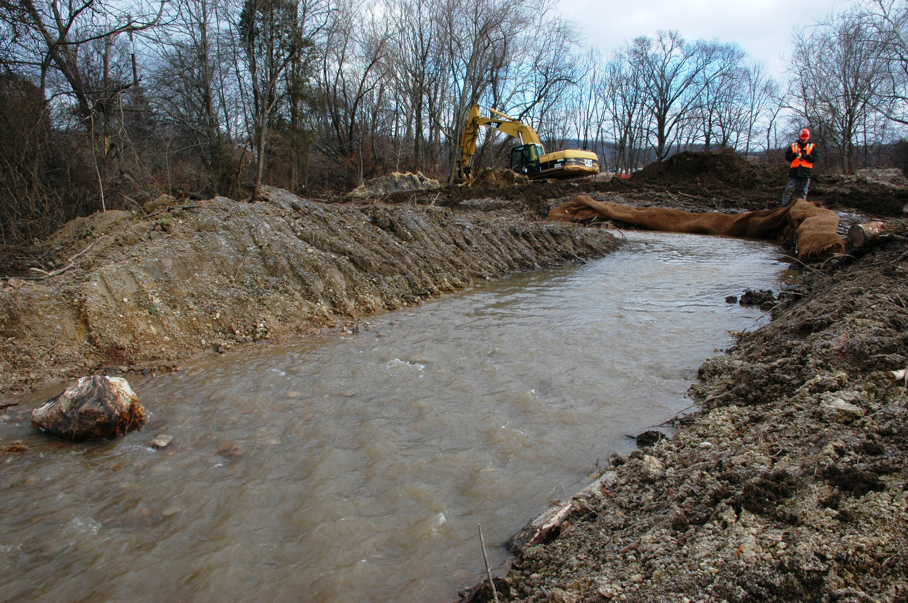 A stream curves between tall, bare earth banks. Matting is laid over the banks to prevent erosion. A man wearing a hardhat stands on one bank. A yellow backhoe sits on the bank behind him.