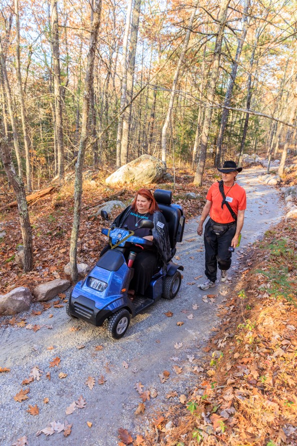 Aislynn Graves rides her mobility scooter on a flat path. Her husband David walks behind her.