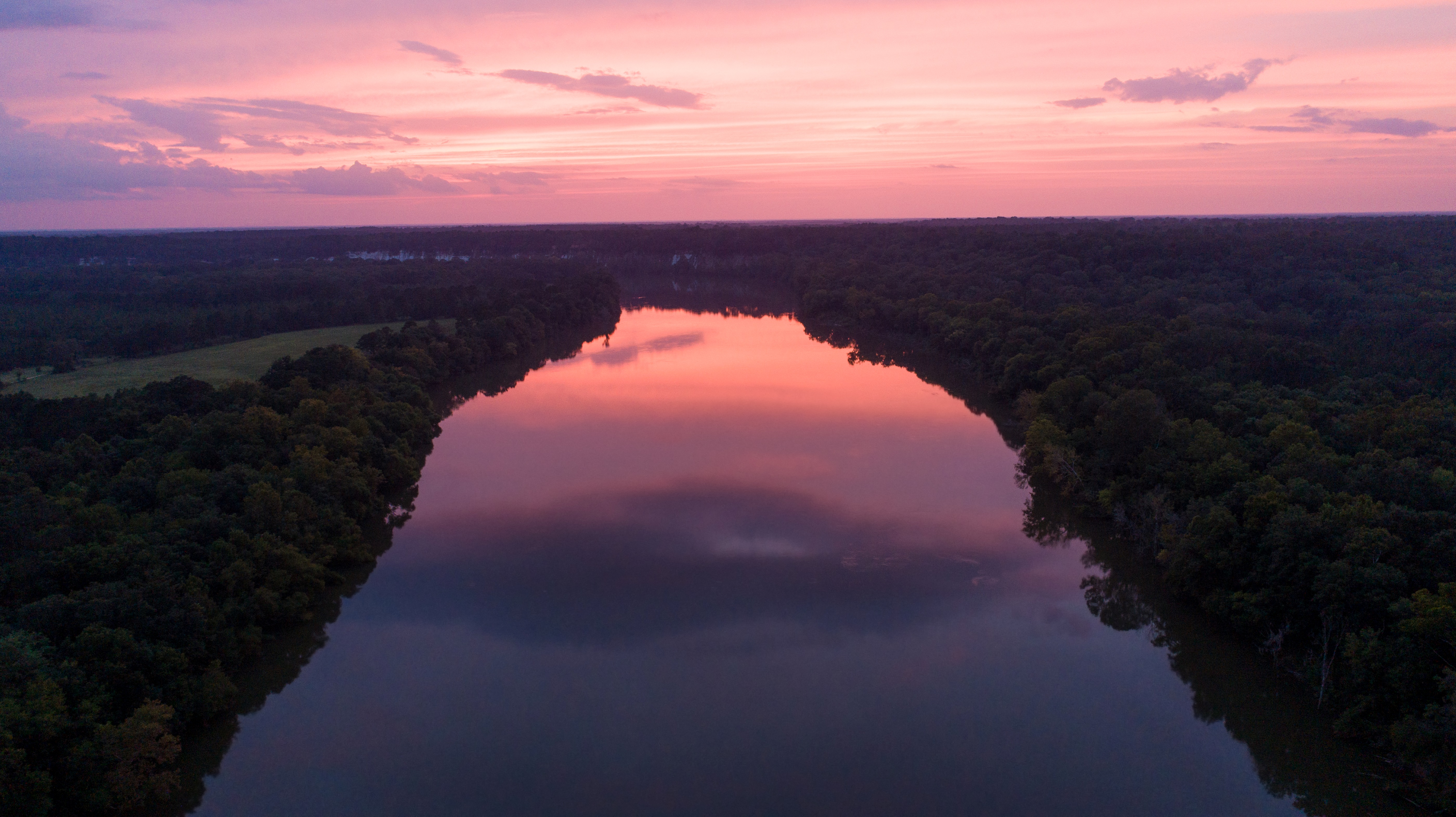 Aerial view of a wide river with thick forests on both banks under a pink sunset sky.