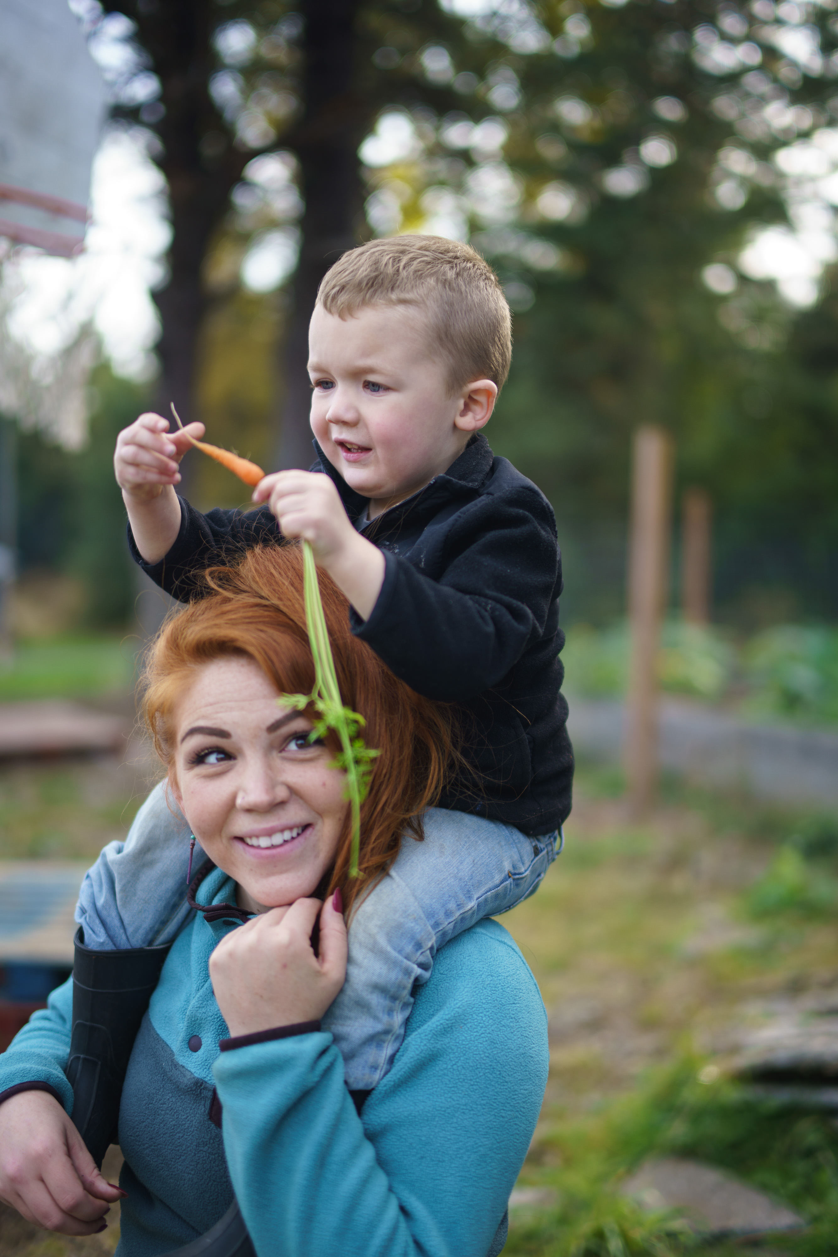 A woman holds a young child on her shoulders; the child holds a carrot in their hands.