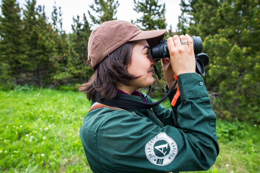 Side profile image of Amanda Cutler holding a pair of binoculars up to her face and looking through them, with greenery in the background.