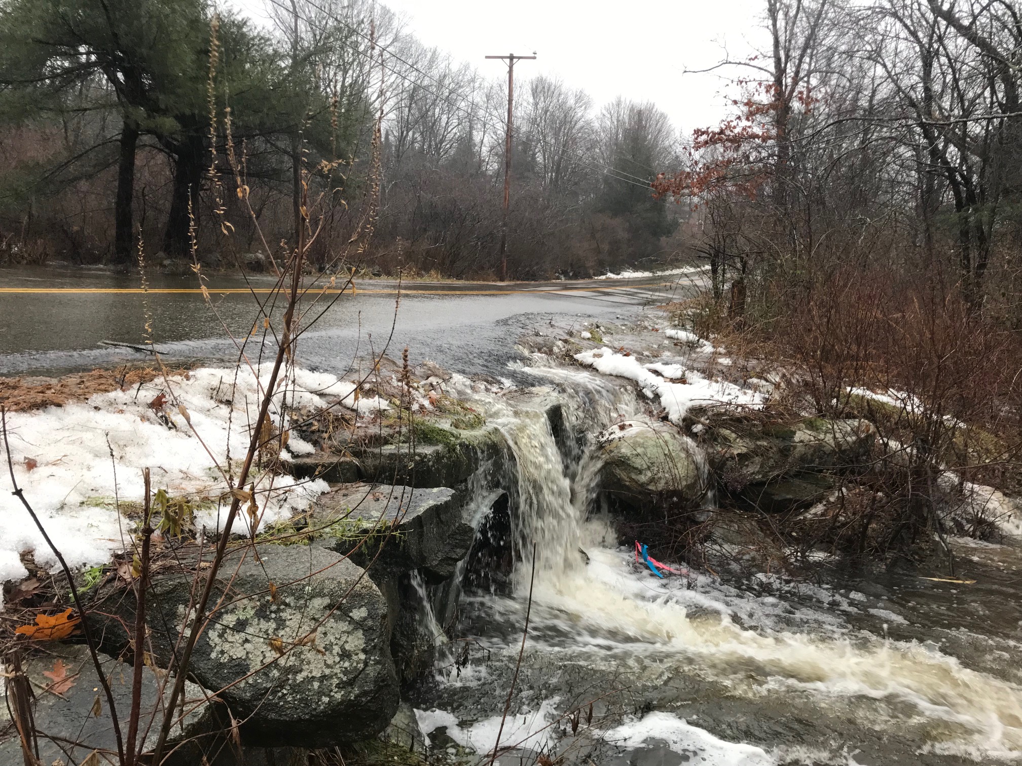 Water rushing across a road and over the rocks along the side of a road instead of going through the culvert.