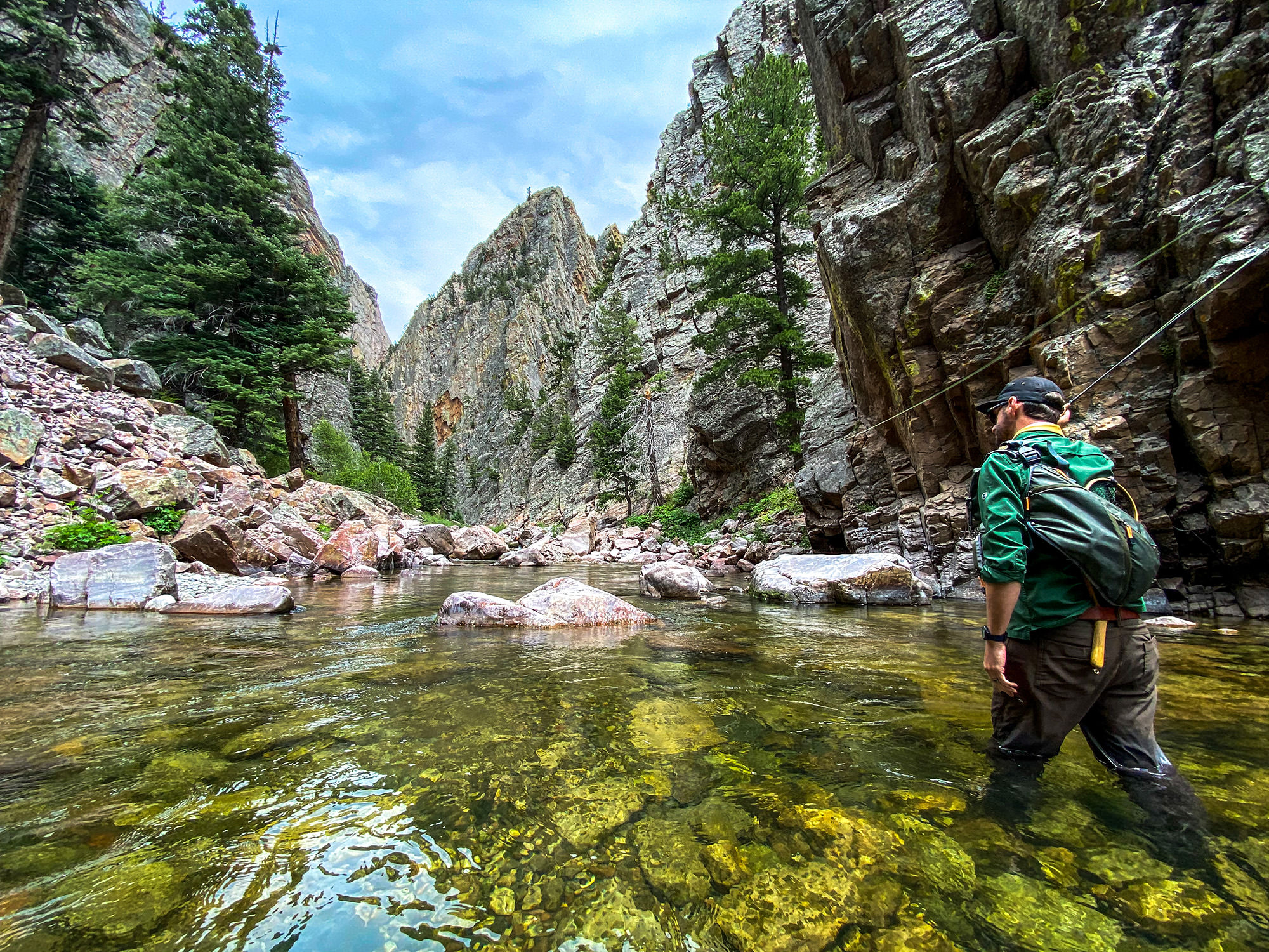 A man wearing a backpack wades through clear water in a rocky canyon.