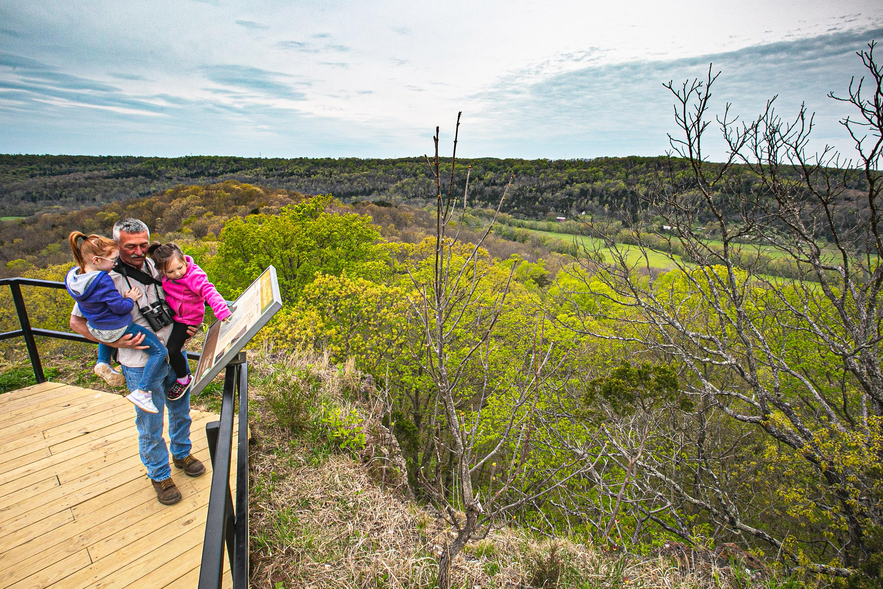 An adult holding two kids reading signage at the peak of Buzzardroot Rock with lush green scenic trees in the backdrop.