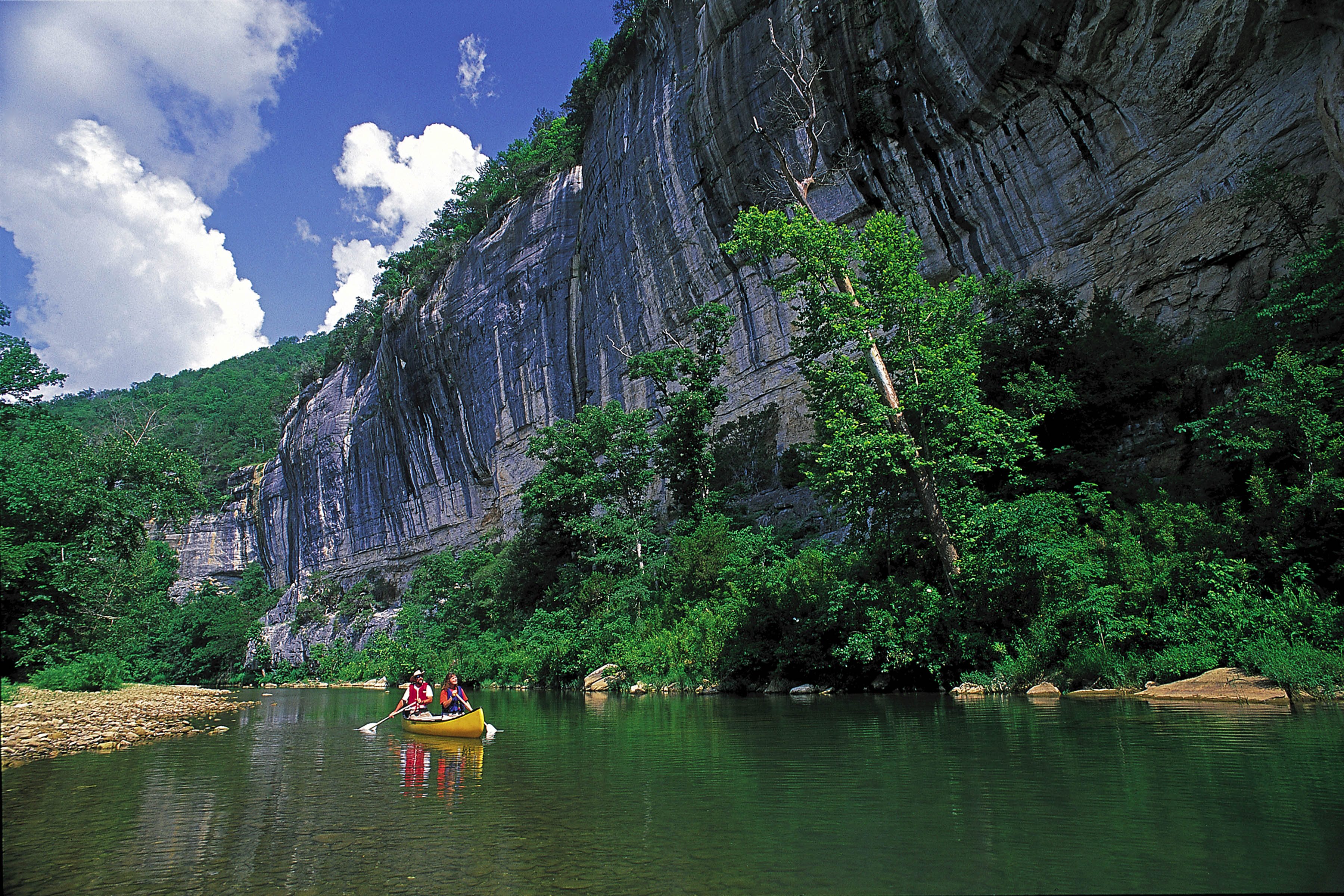 Two people canoeing on the Buffalo River with rocky cliffs along one bank.