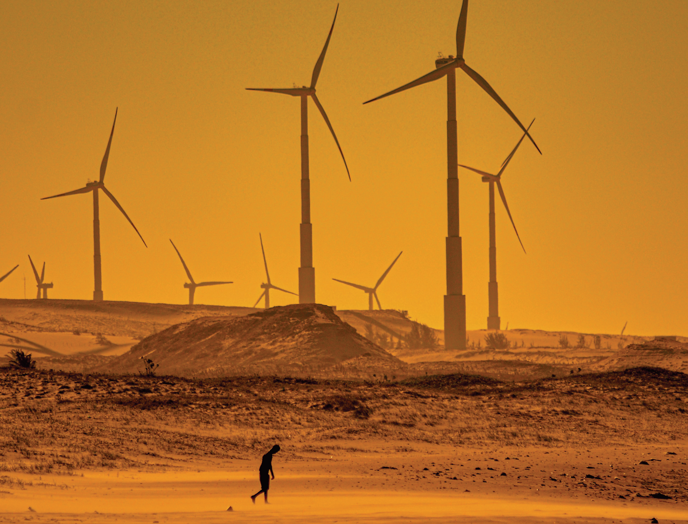 Person walking in desert landscape in front of wind power generators.