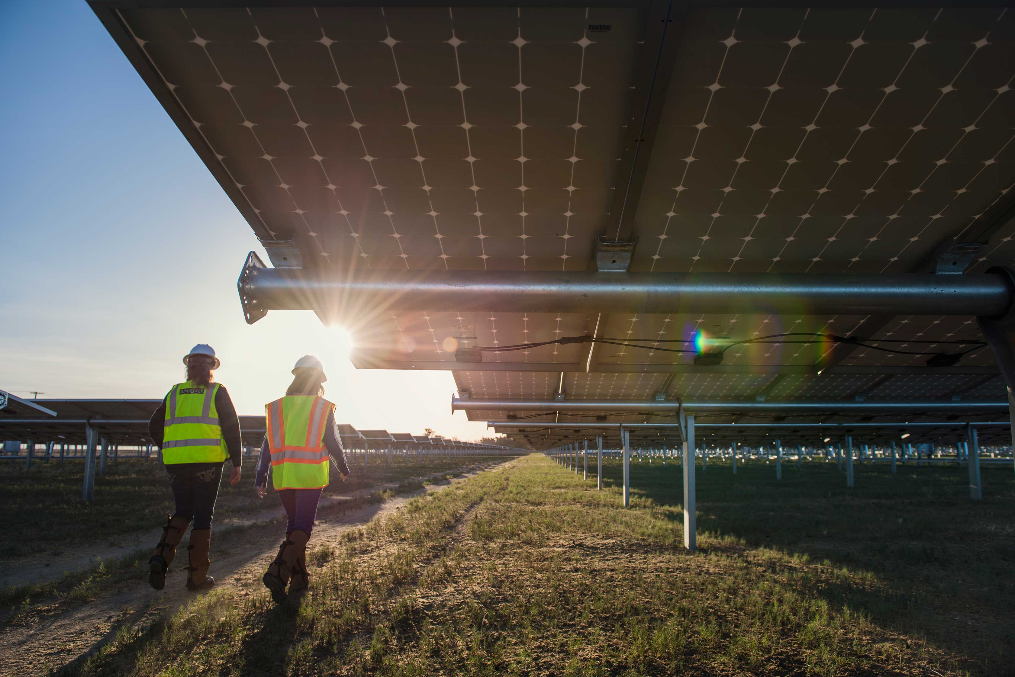 Ground-level view of two women wearing bright yellow and orange safety vests walk next to an array of solar panels.