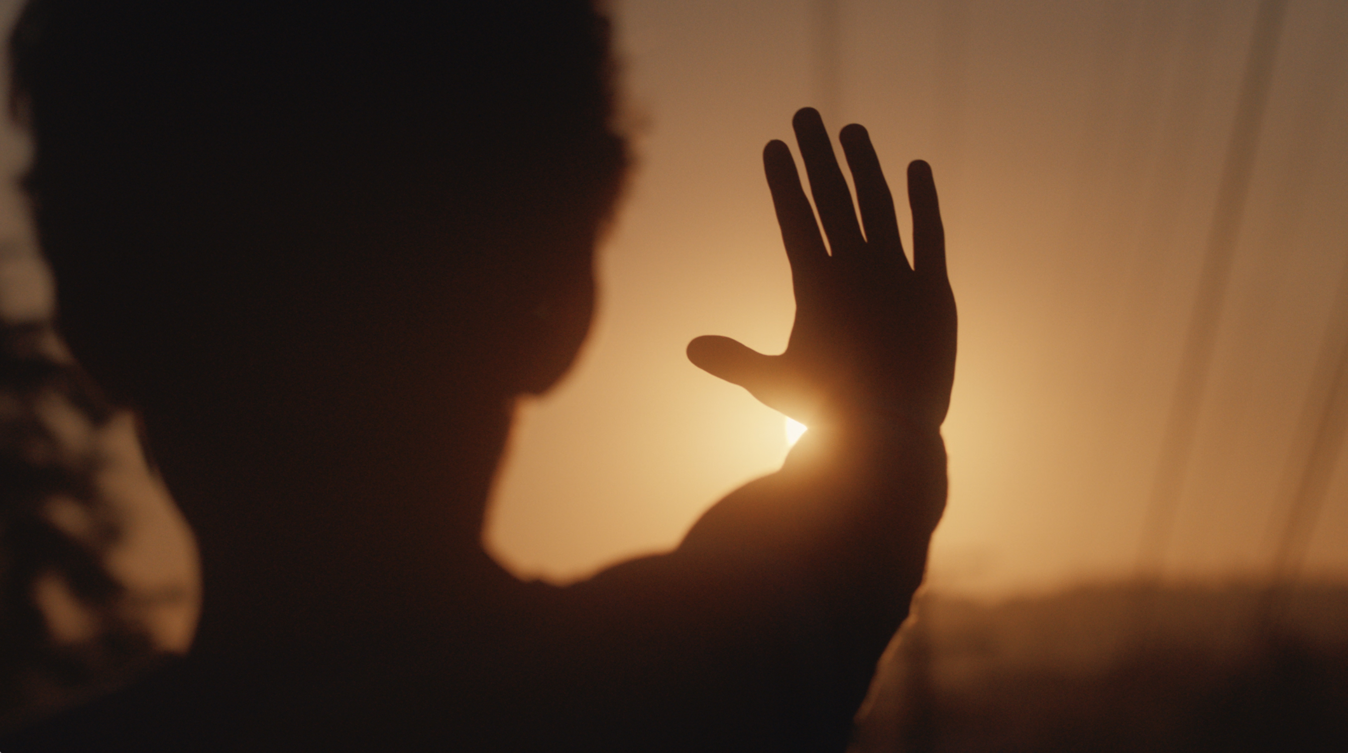 A silhouetted person holds their hand up to block the sun while the sky glows orange.