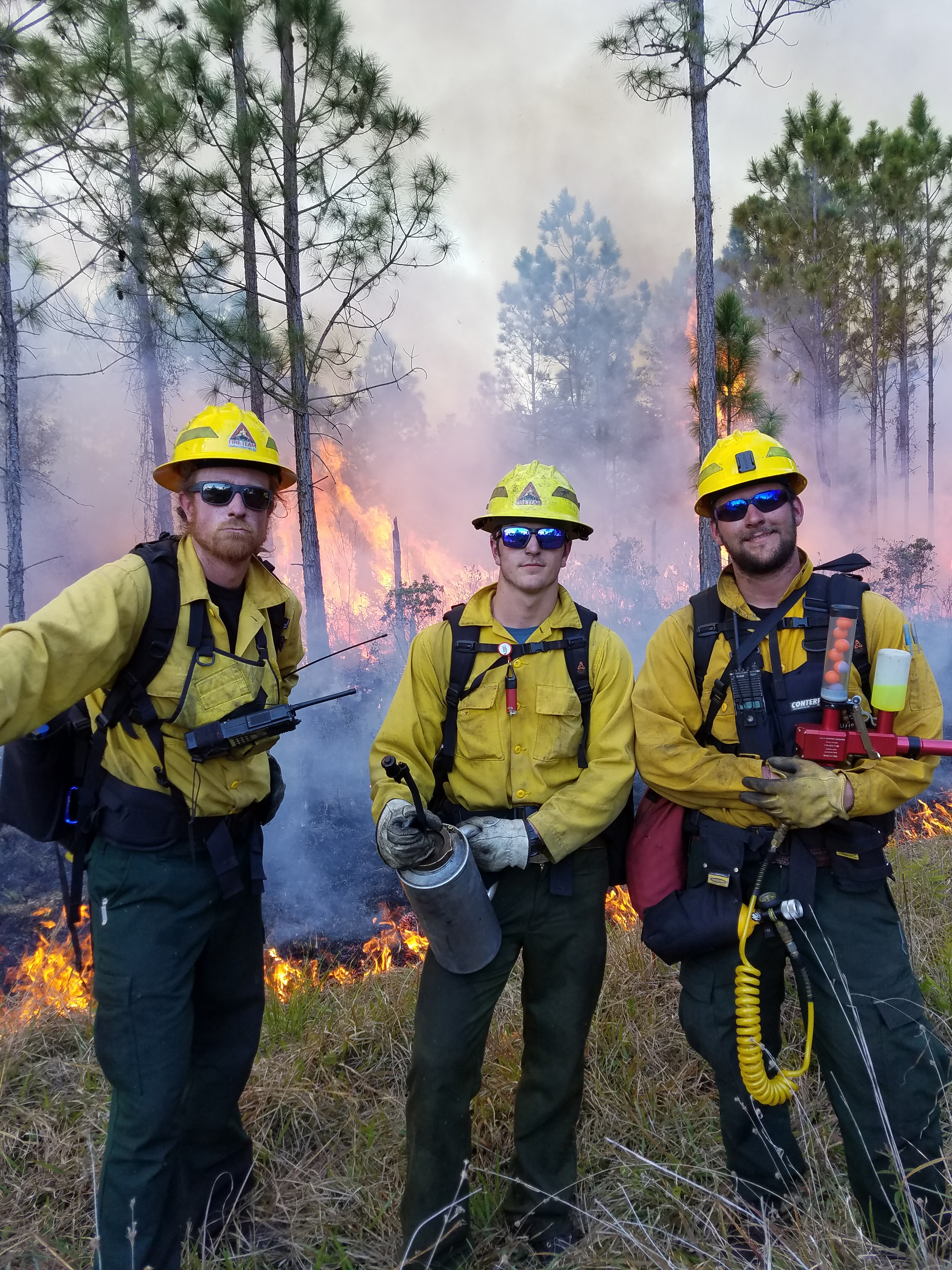 Three students from the University of Idaho dressed in fire-protective gear pose near a prescribed fire at Tiger Creek Preserve. 