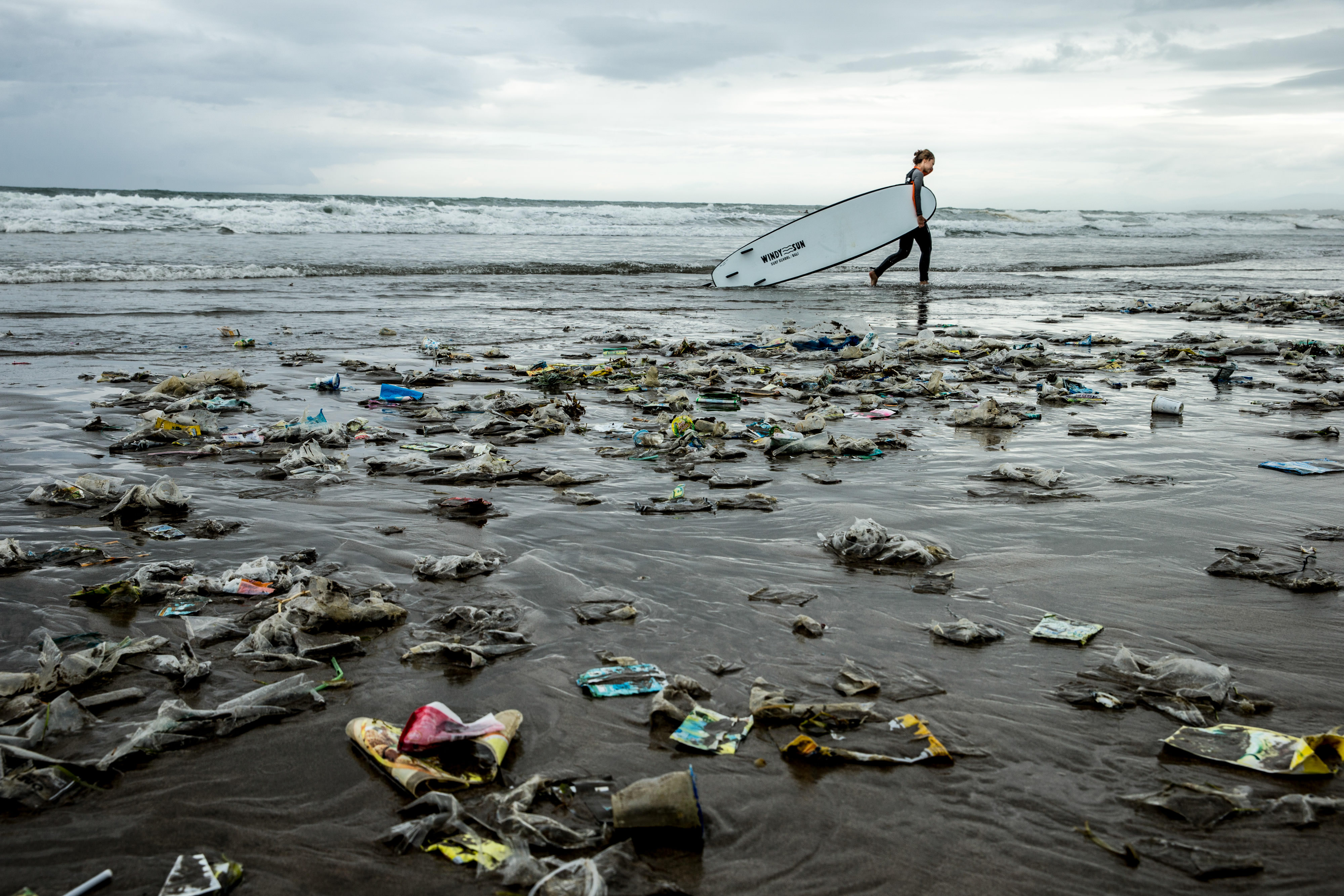 A person carrying a surfboard walks along a beach that is strewn with trash and plastic.