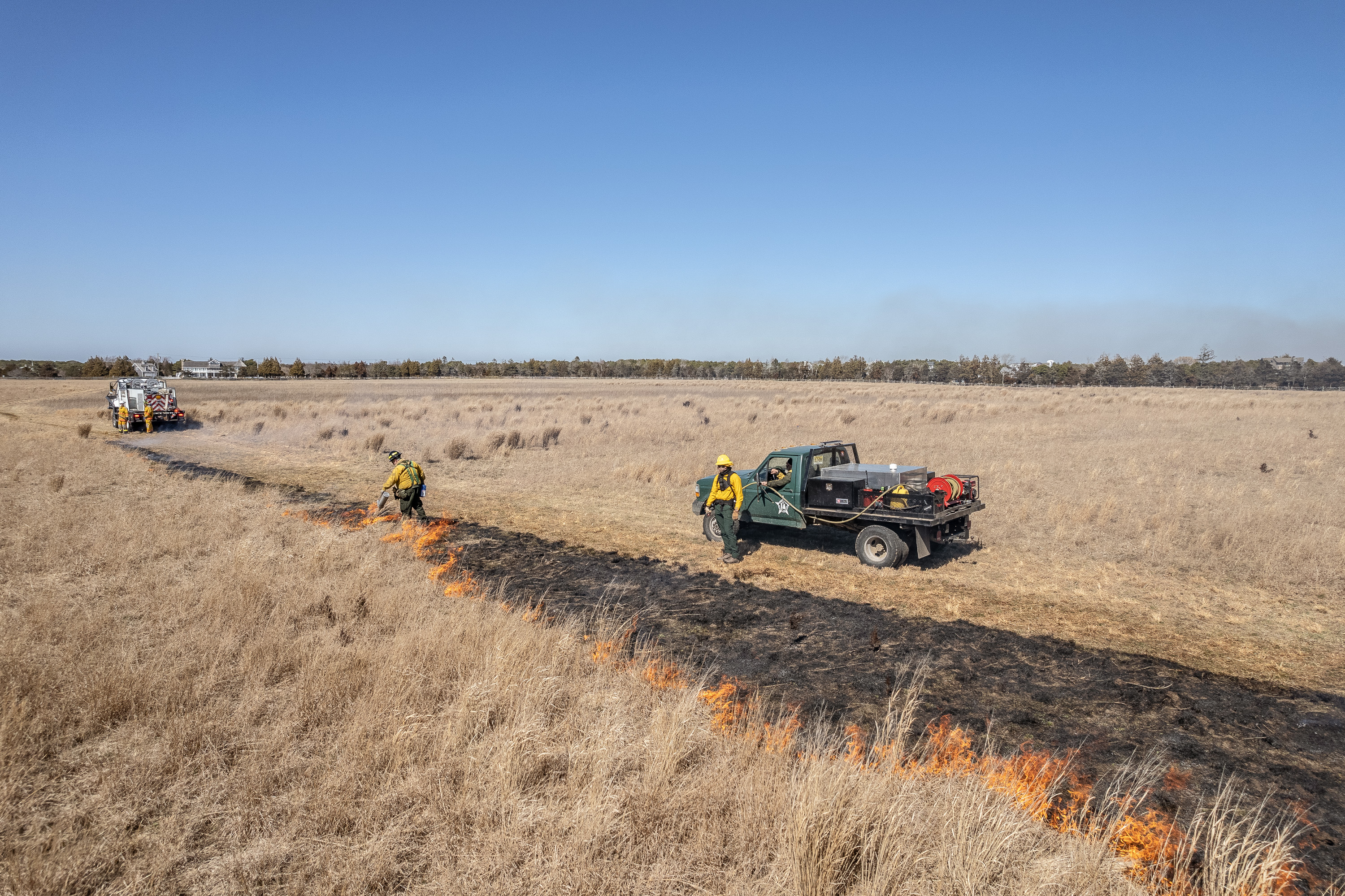 Aerial shot of a prescribed burn crew starting a line of fire through the sandplain grassland, with trucks stationed for support.