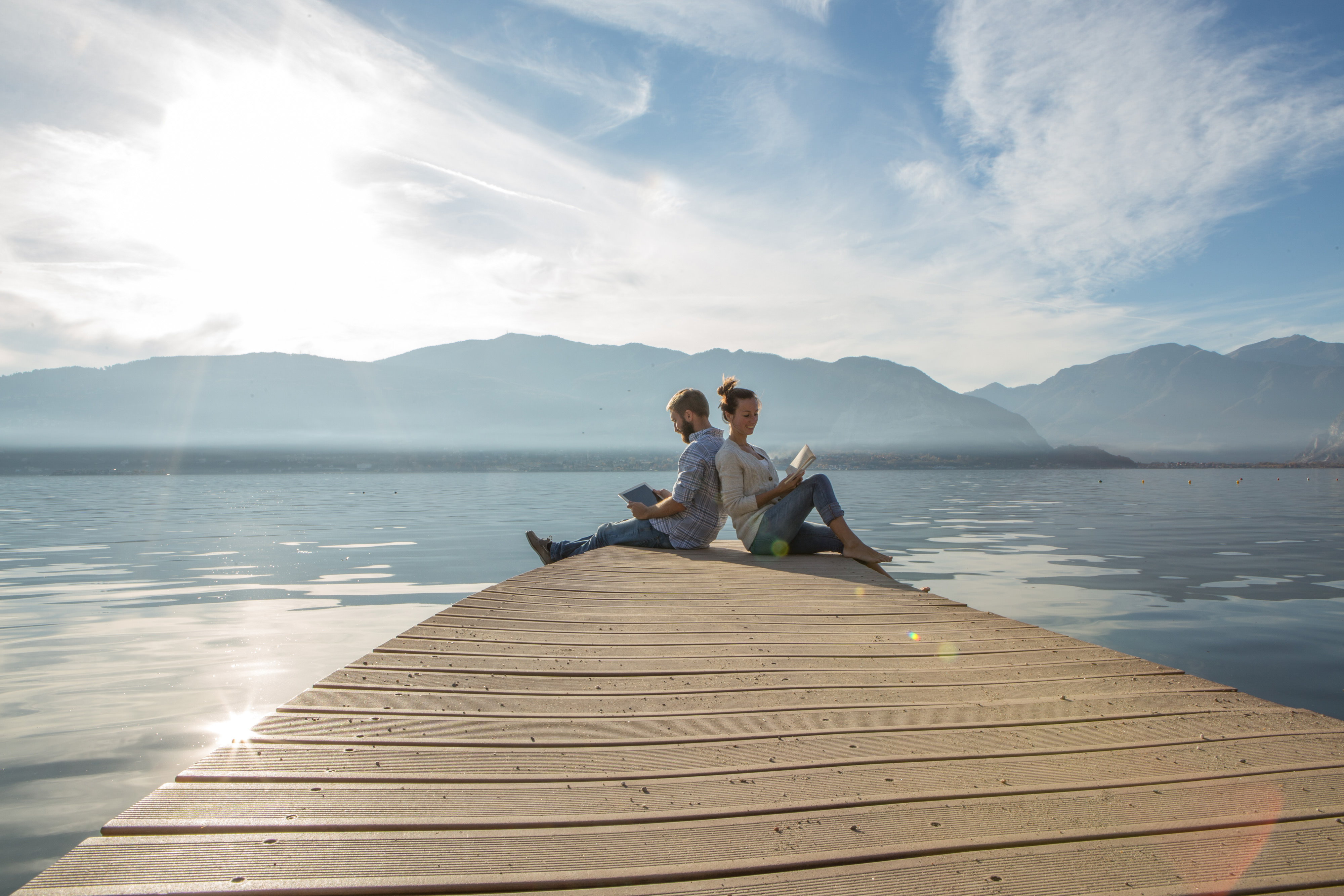 A man and a woman sit back to back at the end of a pier. They're each reading a book. The water behind them stretches to the horizon where it meets a mountain ridge.