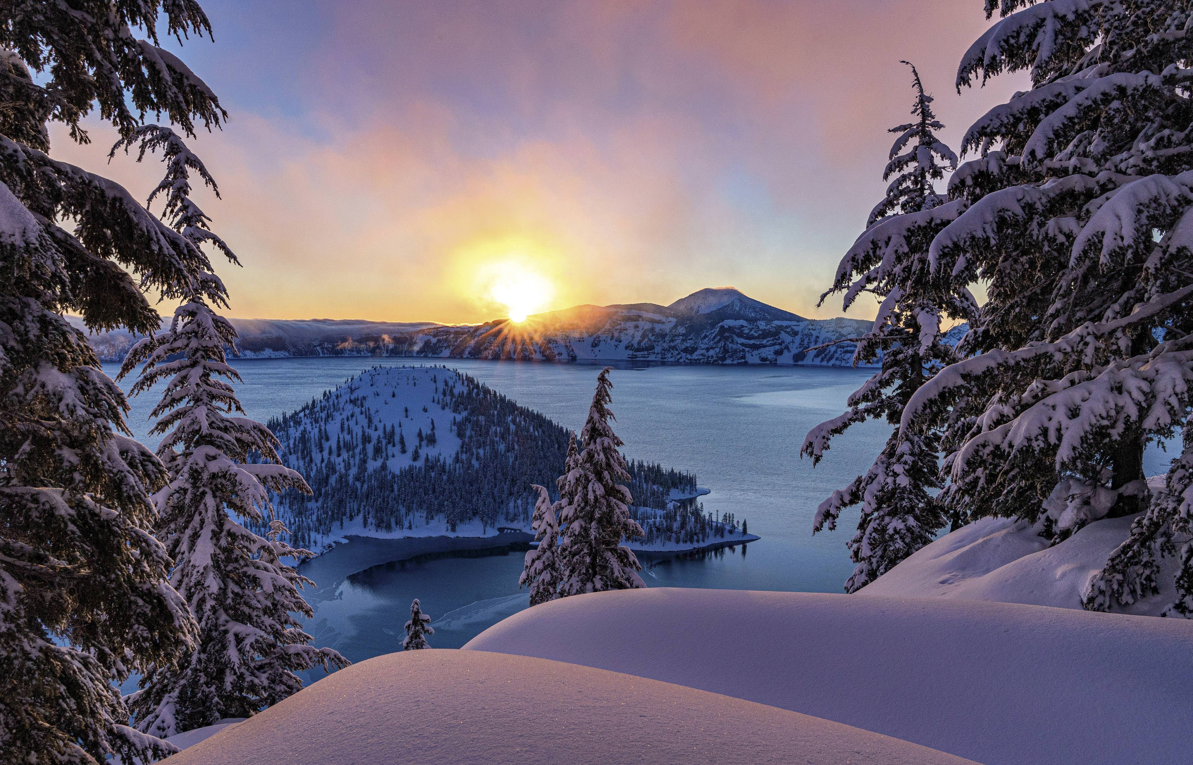 The sun rises in the morning over snowy Crater Lake National Park in Oregon. There's a view of the lake and Wizard Island.