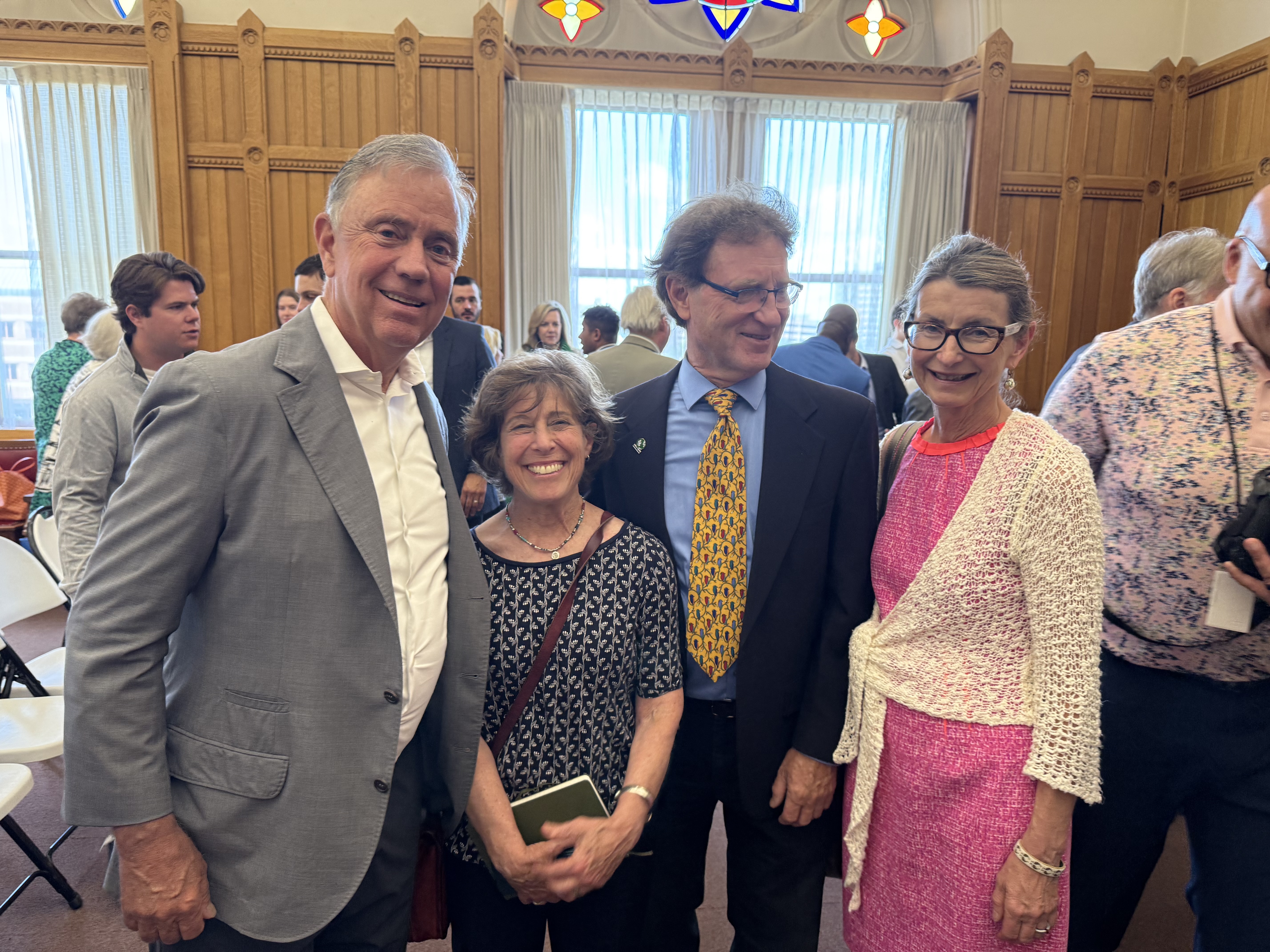 A group of four people is standing together, smiling in a room with large windows and wooden paneling. They appear to be at an event or gathering.
