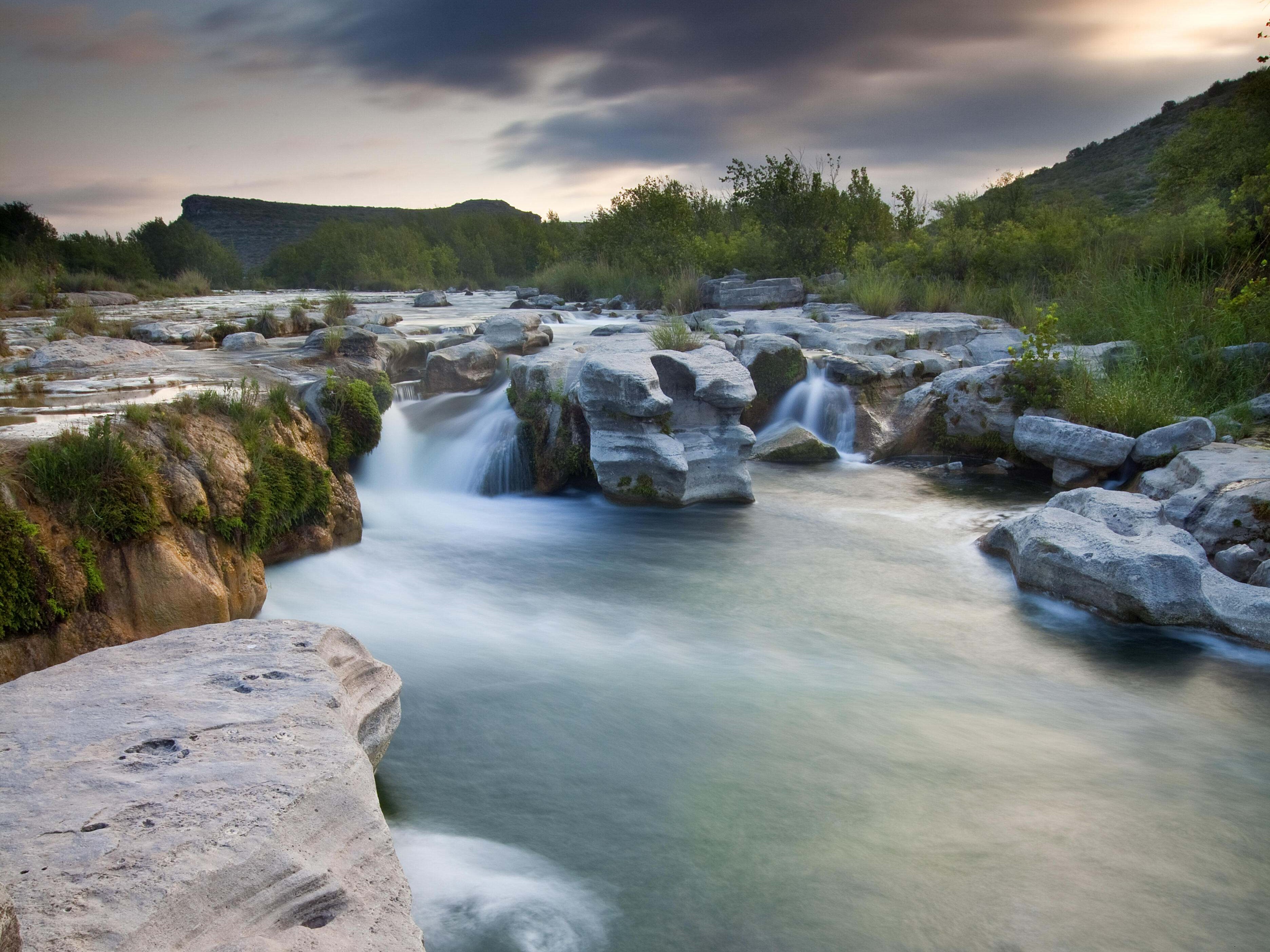 Turquoise waters flow and fall over stacked rocks surrounded by greenery.