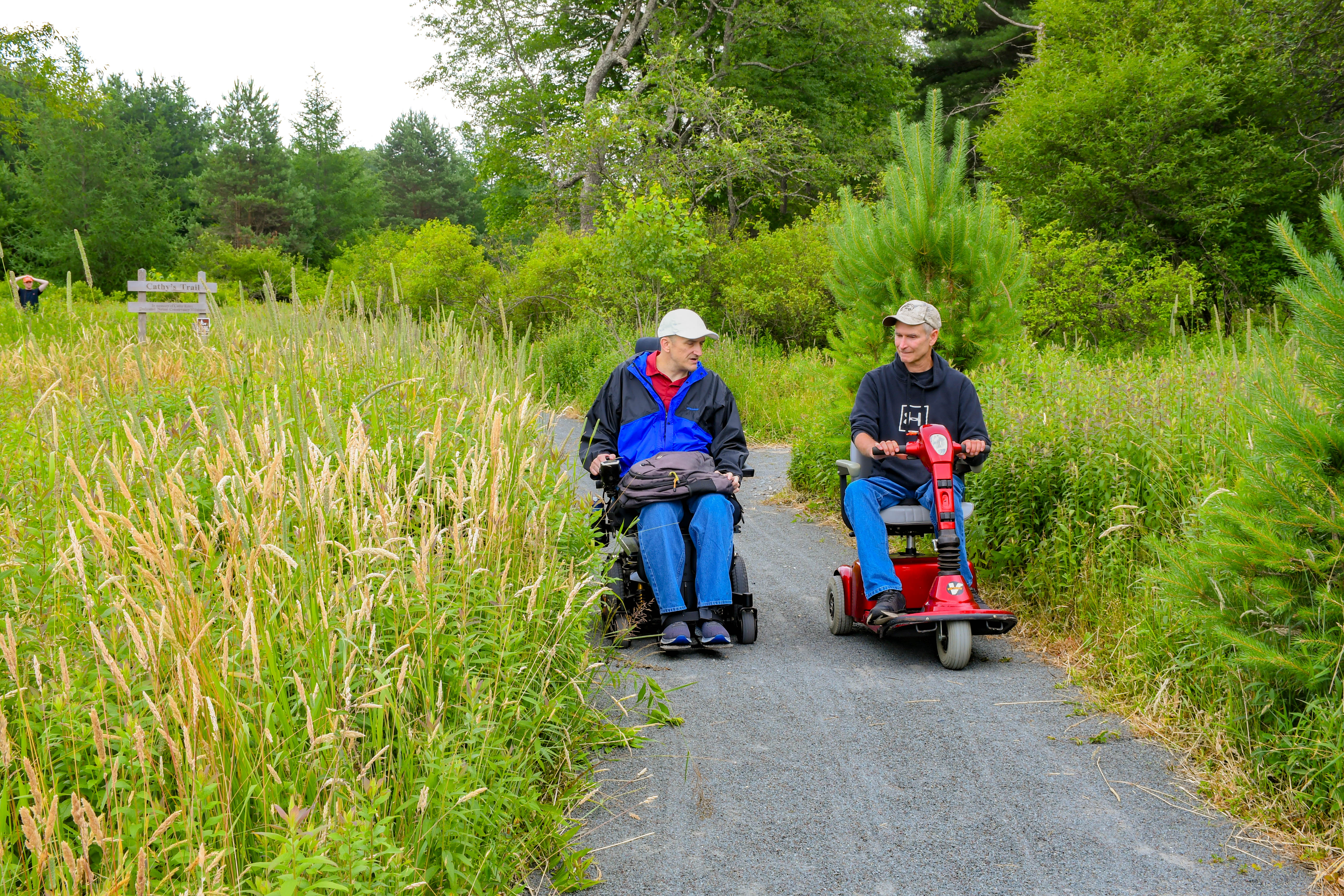 Two people using mobility devices enjoy a paved trail.