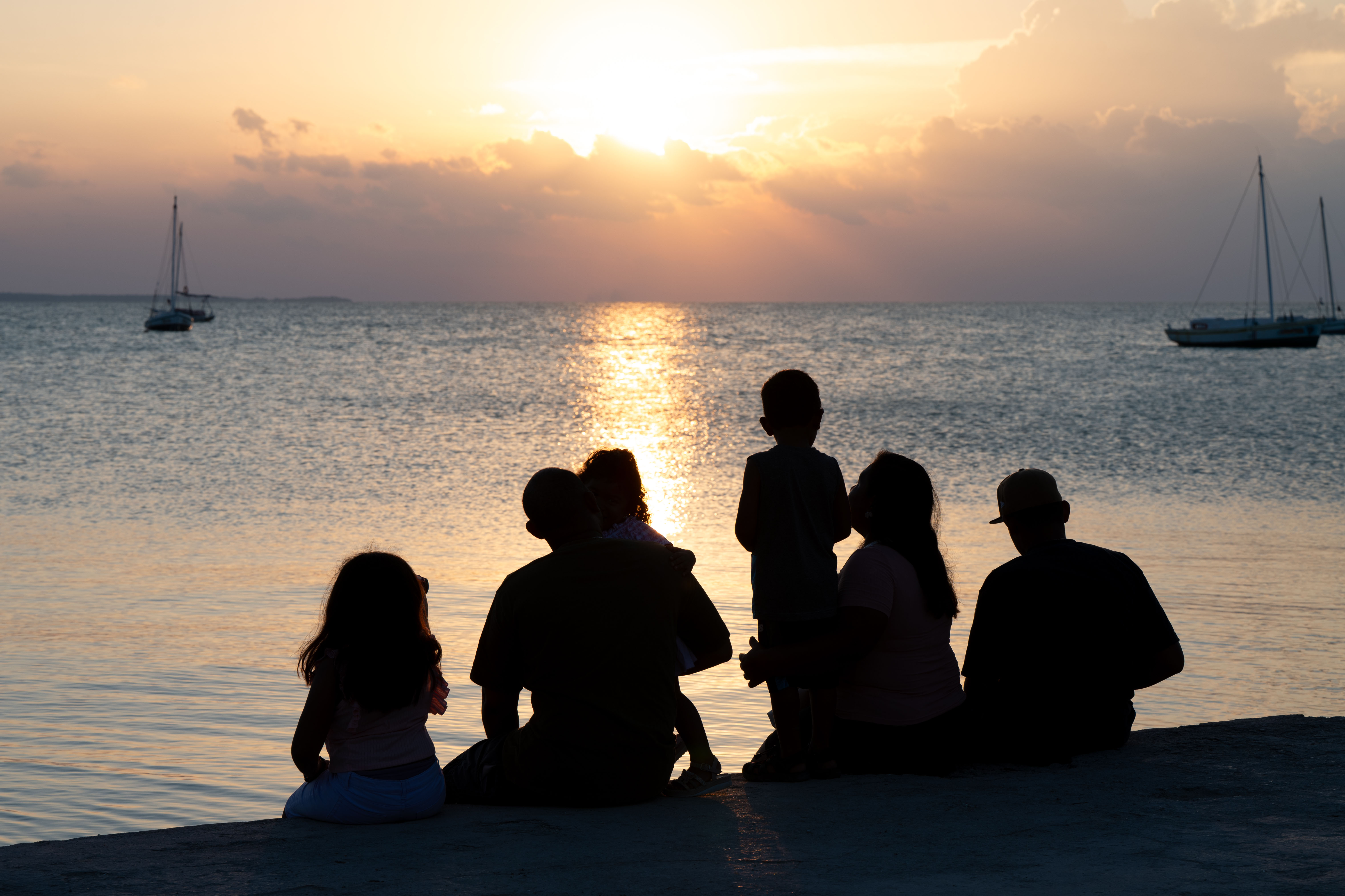 A family of several adults and children are silhouetted as they sit on a beach and watch the sun set over the ocean.