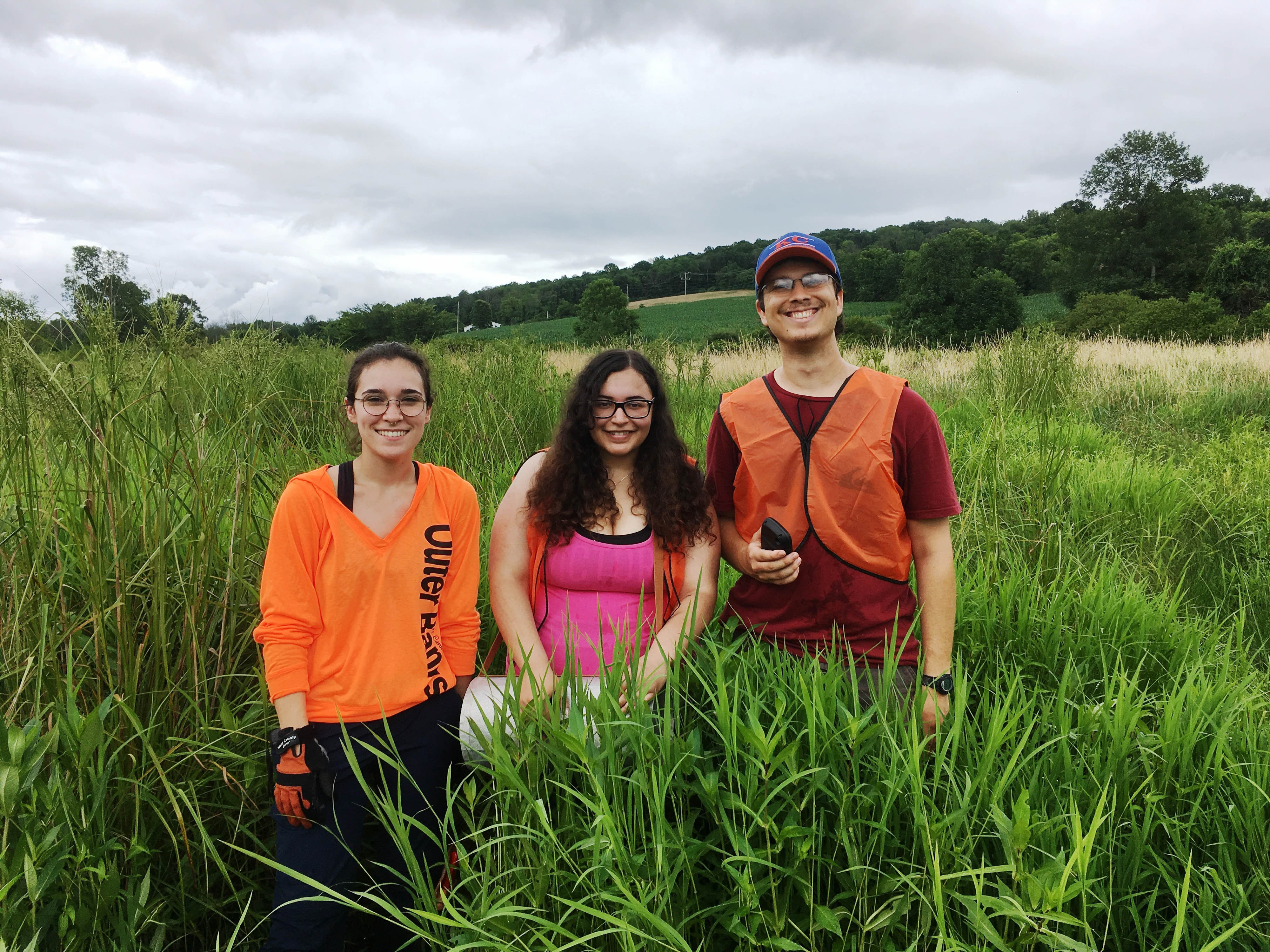 Three people, two women and a man, stand together in waist high vegetation, posing and smiling. The roof of a green building is visible behind them. A mountain ridge runs along the horizon.