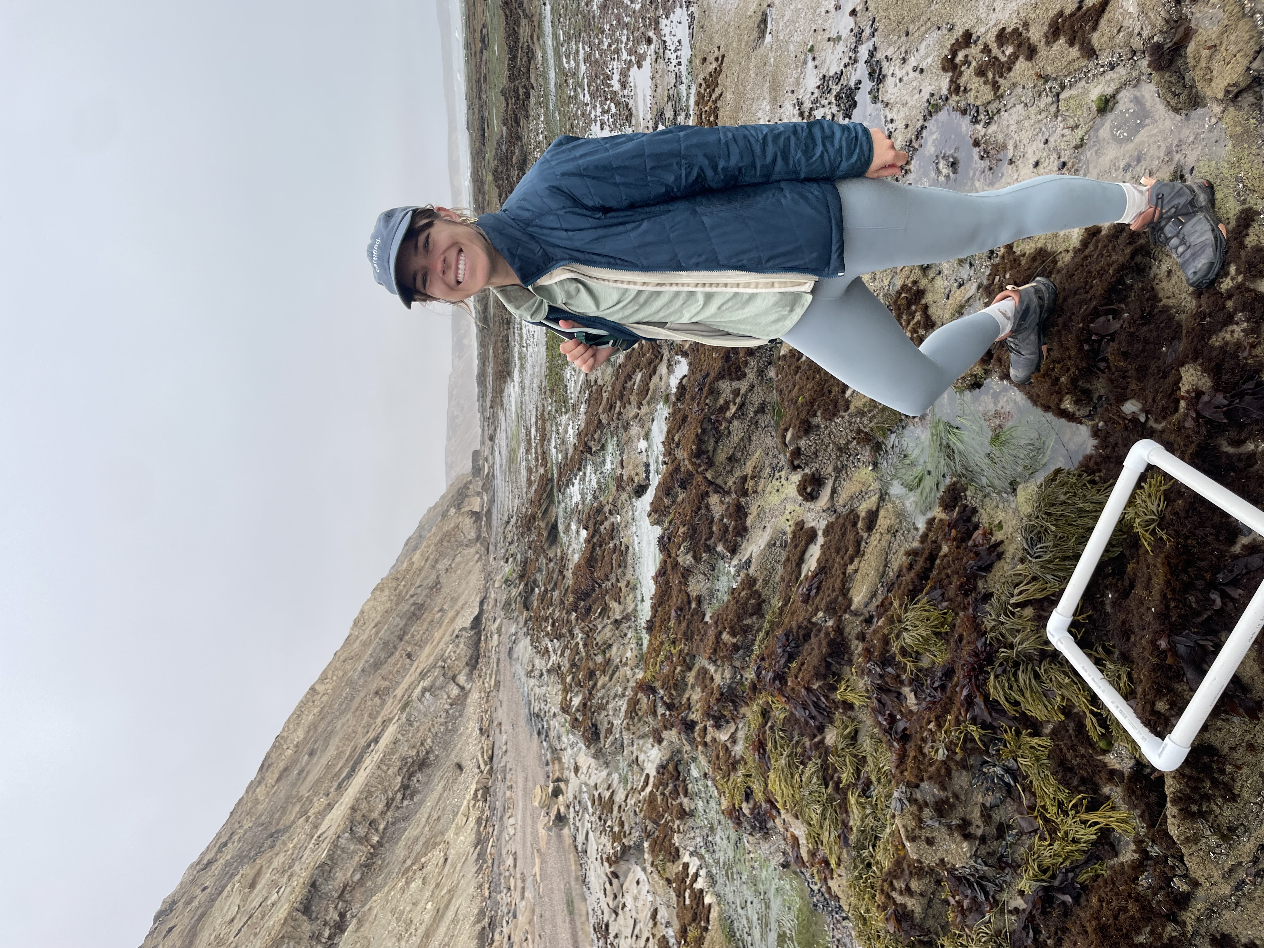 A woman wearing a jacket and hat poses along a coastal area.