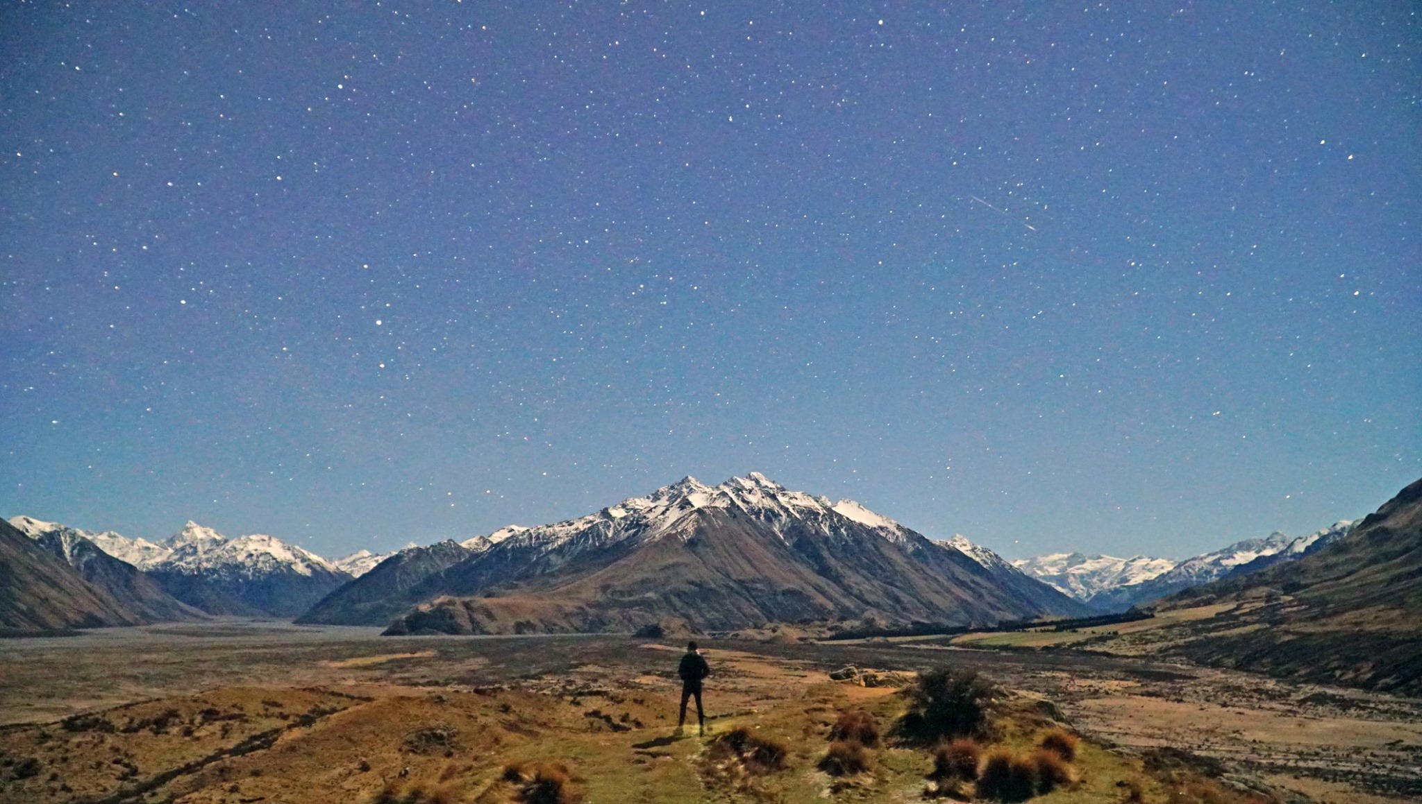 man looks at snowcapped mountains, grassland and stars