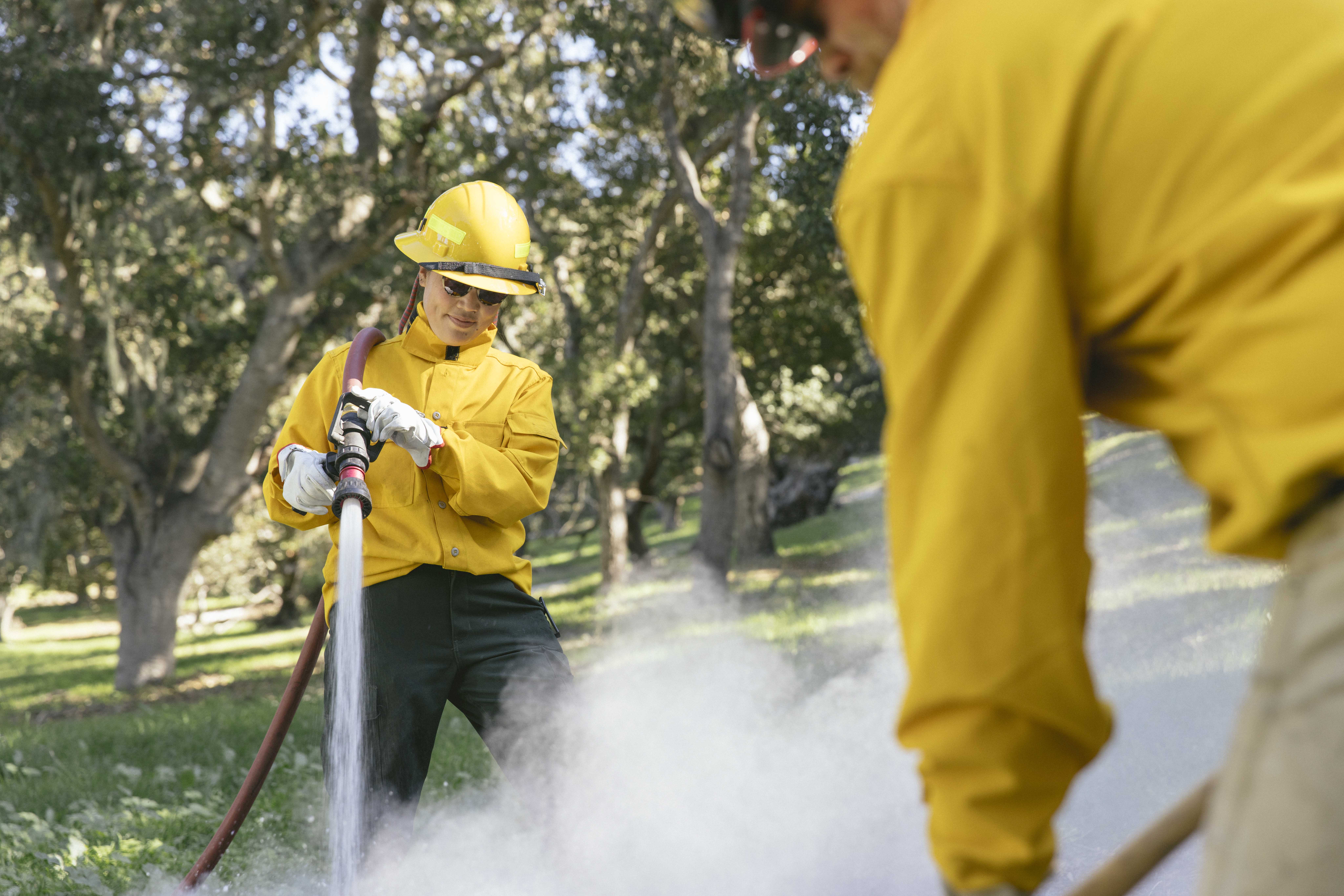 A woman in a yellow jacket holds a fire hose over her shoulder while she soaks ashes after a controlled burn.