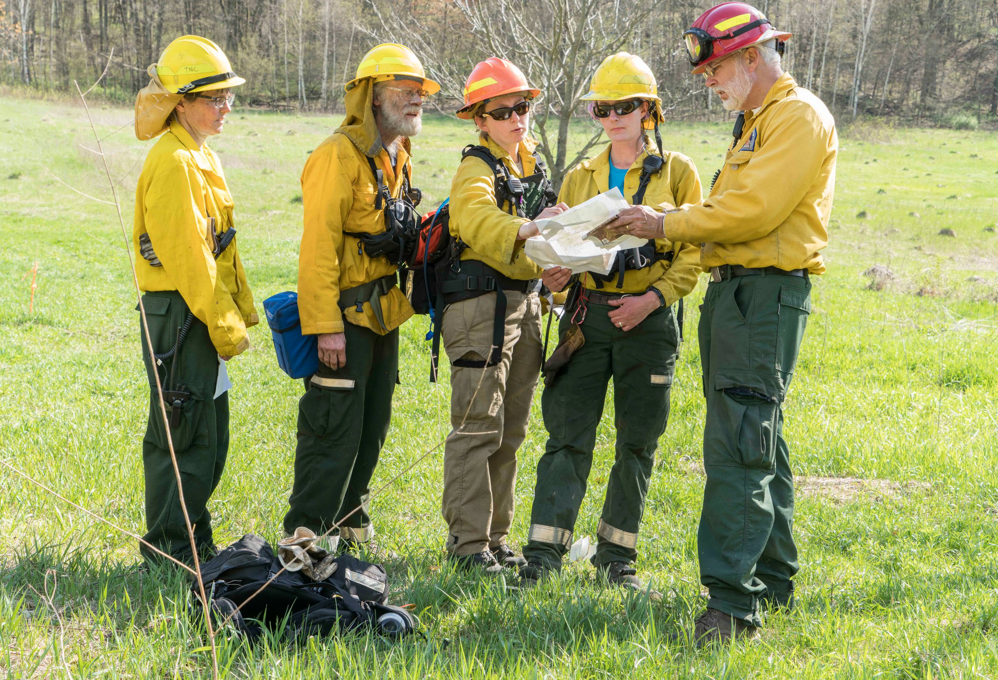 Five people dressed in yellow Nomex suits, helmets, goggles and other fire gear consult maps while working on a prescribed fire in Wisconsin.