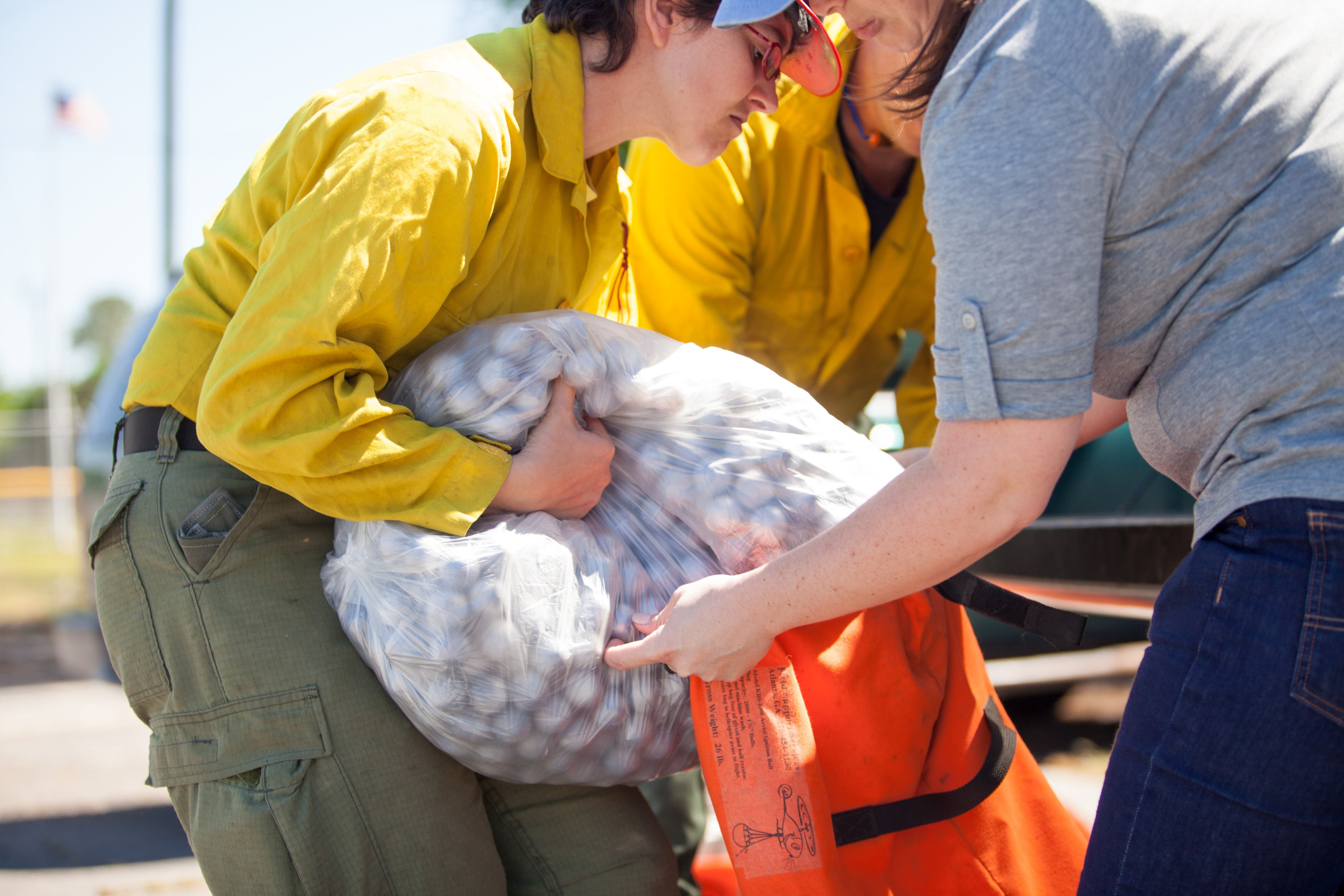 Three people huddle around an orange sack. One person holds the sack open while a second empties a clear plastic bag full of gray spheres into the sack.