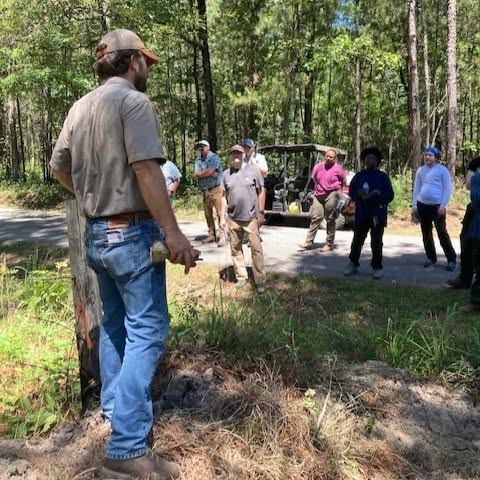 A person talks to a gathering of people near a forest.