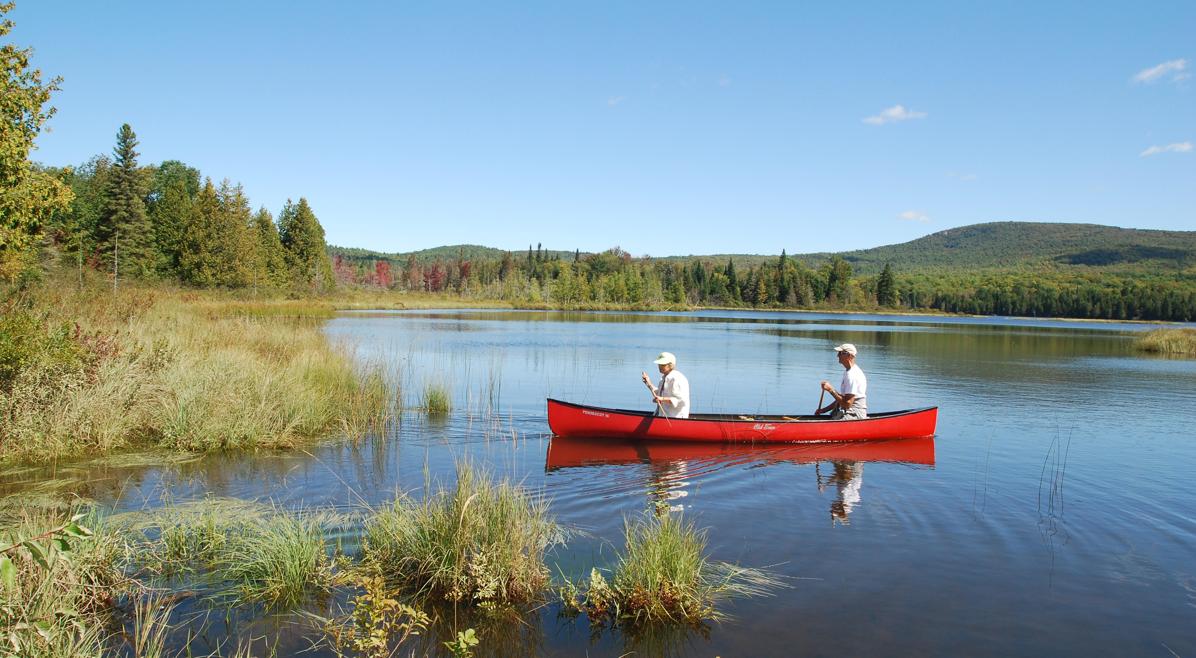 Two people paddle a red canoe on a pond with forested mountain slopes in the background.