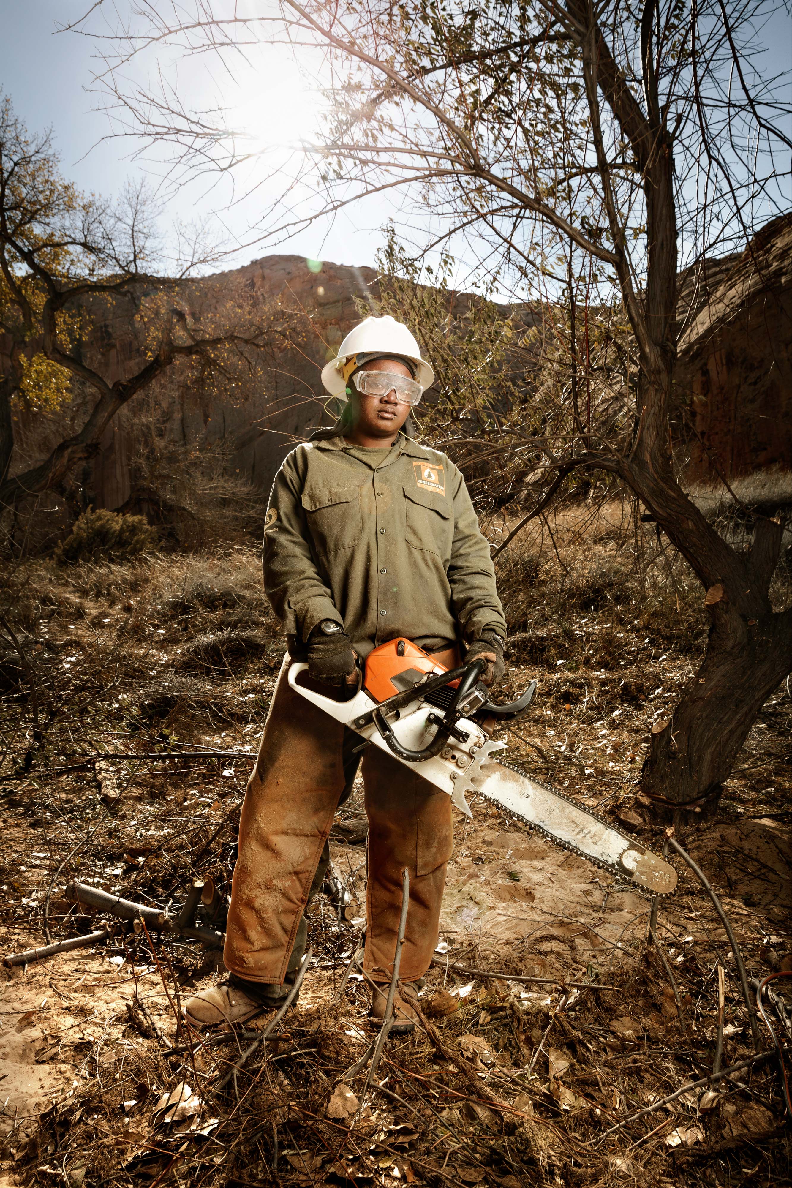 A young black woman in protective clothing with safety glasses and helmet, holding a chainsaw and looking at the camera.