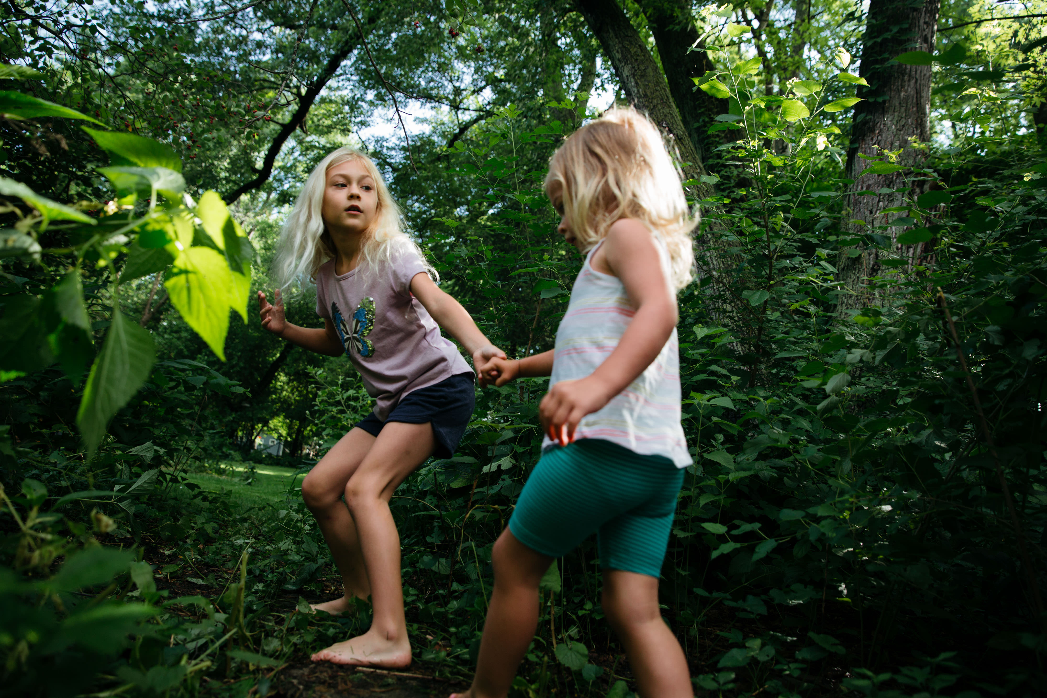 A barefoot girl reaches for the hand of a younger also barefoot girl to draw her through a gap in some bushes.