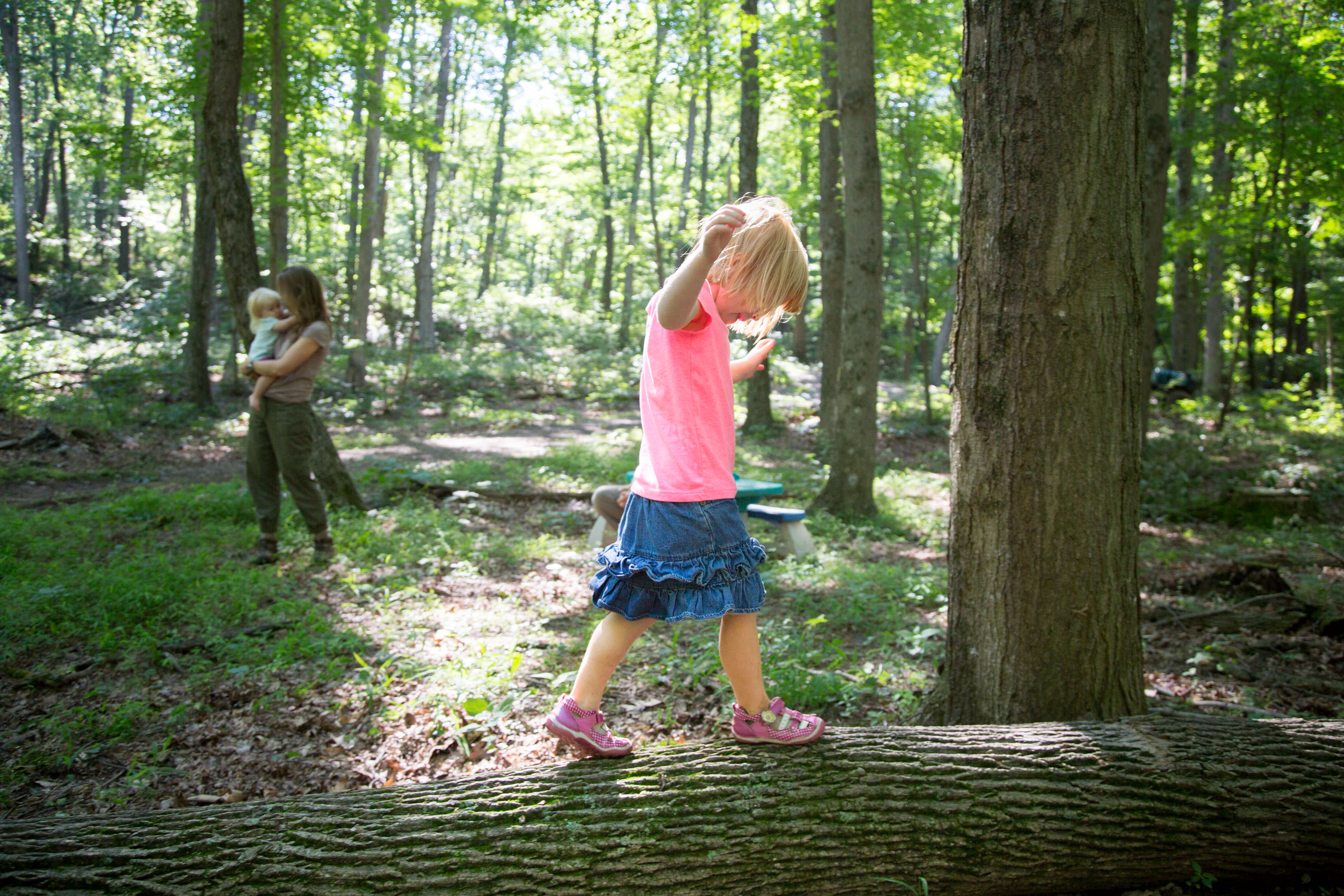 A young girl wearing a pink top and blue skirt balances on a fallen log in the middle of a sun dappled forest.