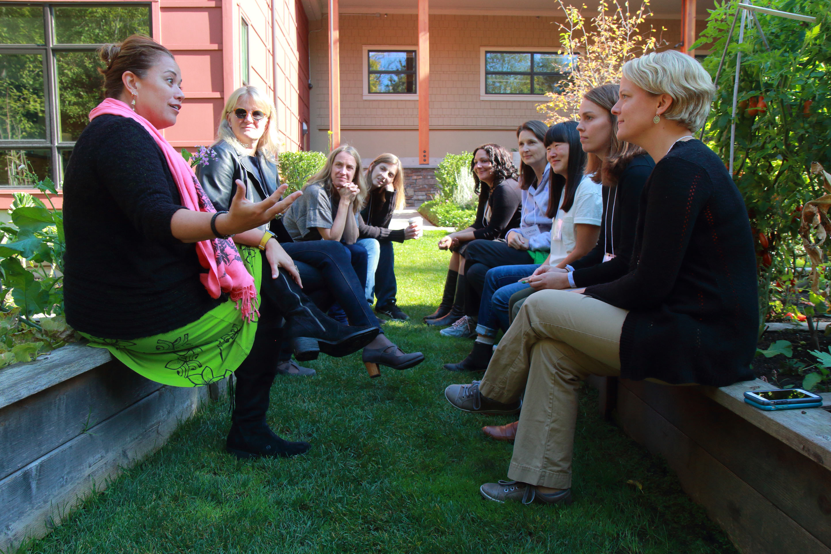 A group of women sit across from each other on benches in a grassy area outside a building. 