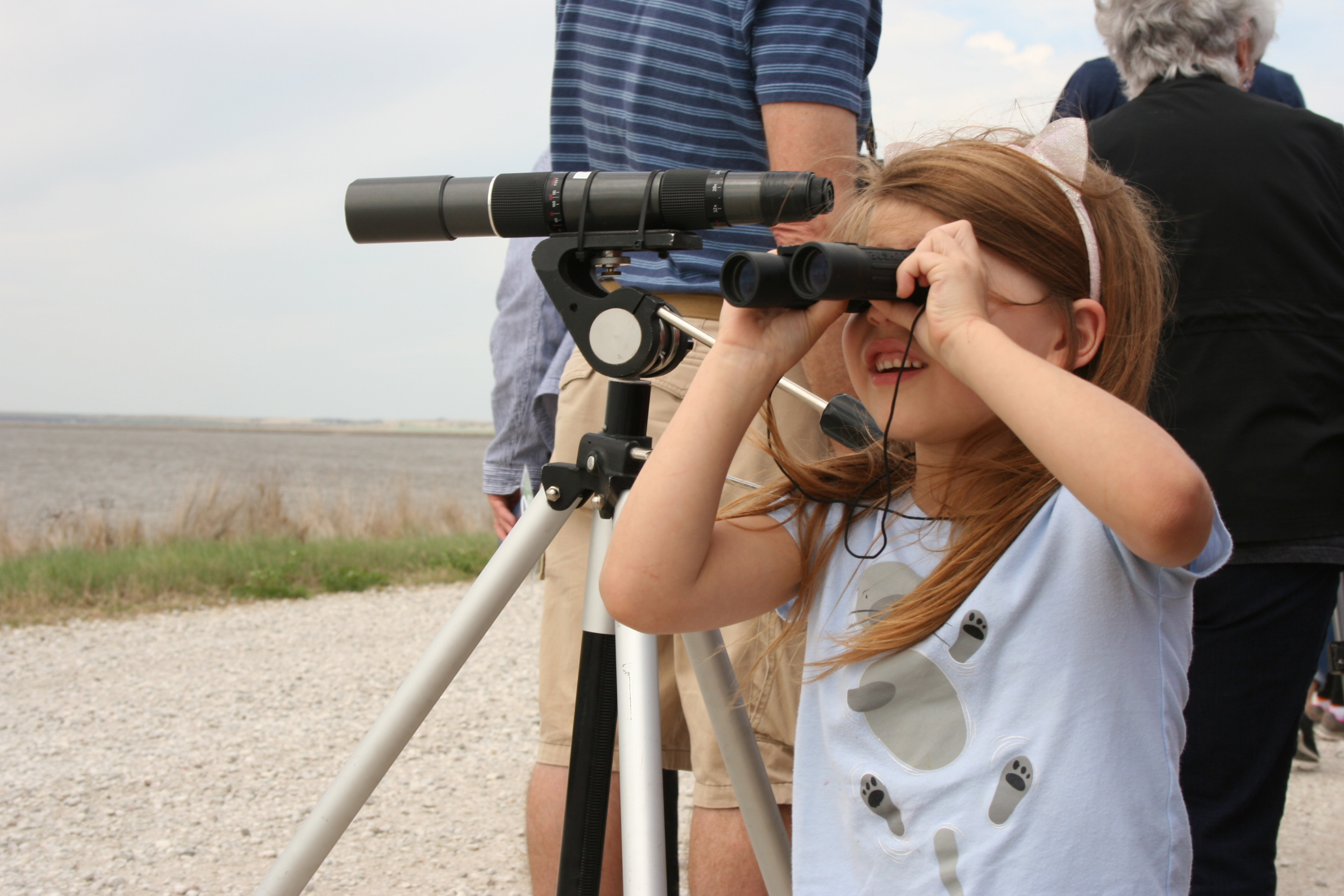 A little girl looks through bionoculars while standing next to a sighting scope.