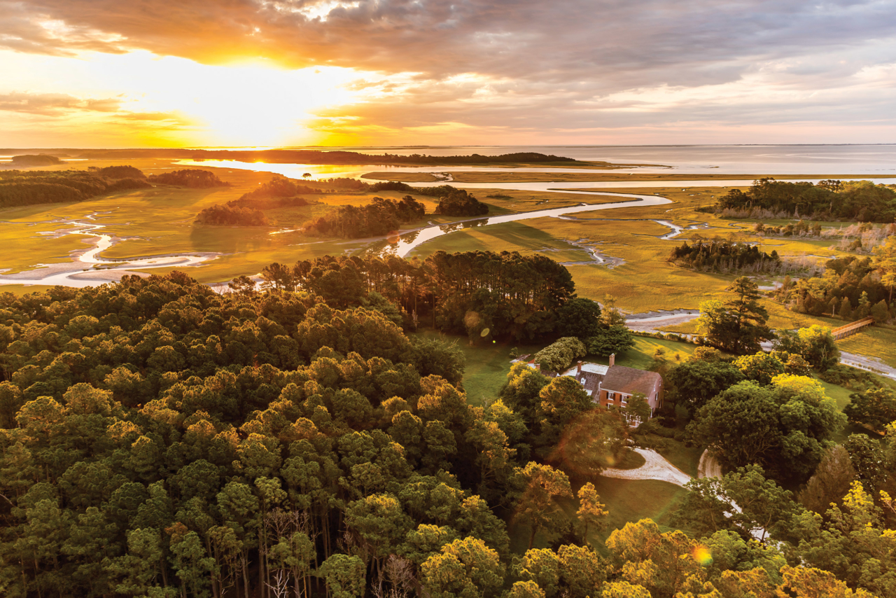 Aerial view of Brownsville Preserve. An historic brick home with a circular oyster shell driveway stands in a clearing between thick stands of trees. In the background the sun rises over the Atlantic.