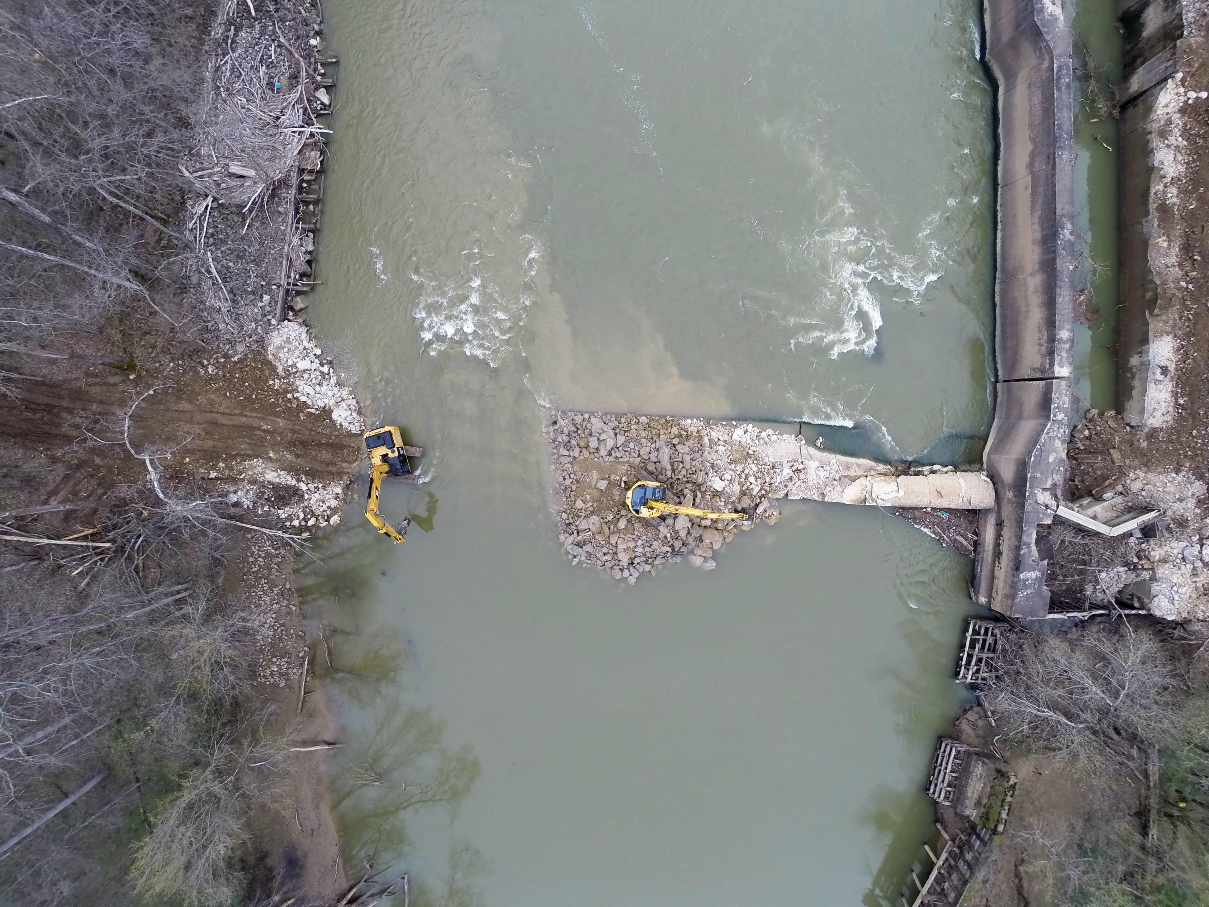 Aerial image of dam removal, looking down on diggers that have partially removed a concrete dam from the river.